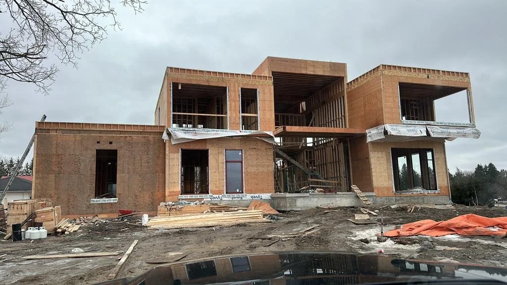 A two-story house under construction with exposed wooden framing and brick exterior, surrounded by construction materials and debris, on an overcast day.