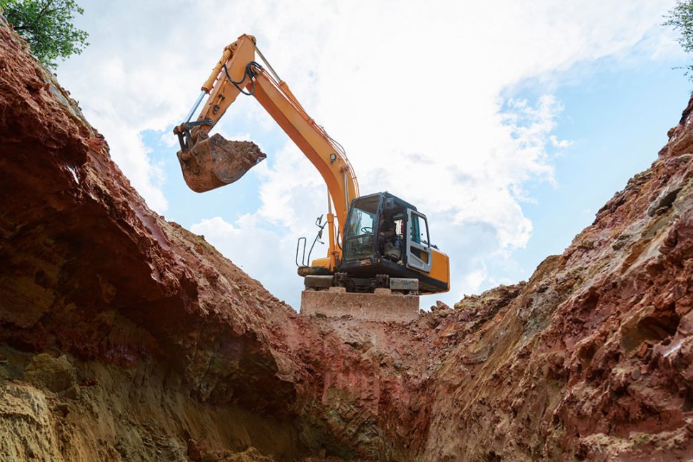 An excavator with a yellow and black body is digging in a trench, with dirt and soil surrounding it under a cloudy sky.