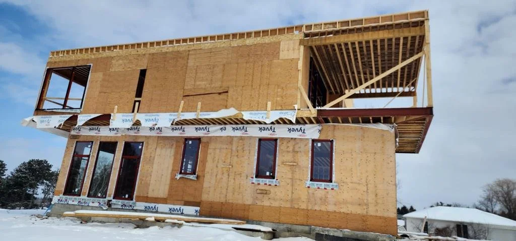 A two-story house under construction with wooden framing and plywood exterior in a snowy landscape.