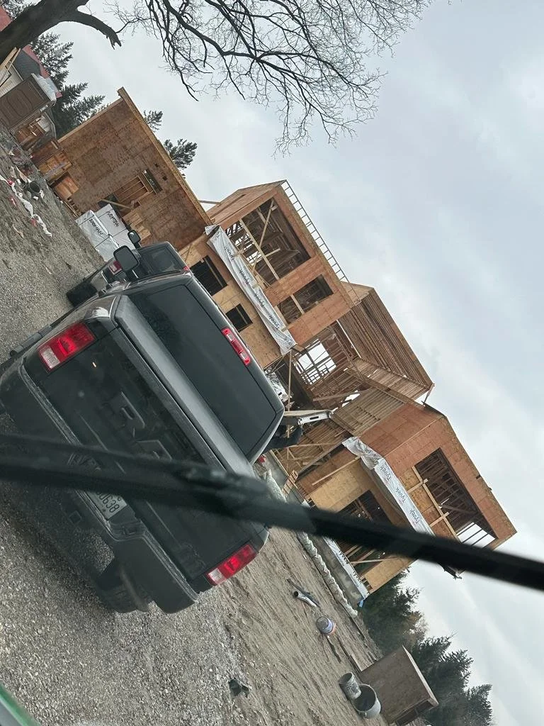 A house under construction with a wooden frame and scaffolding, surrounded by construction vehicles and building materials on a cloudy day.