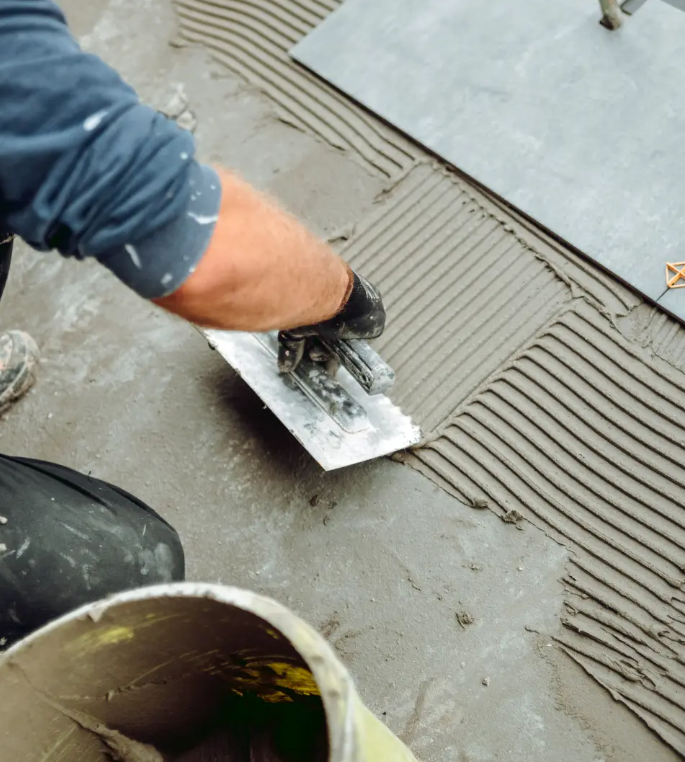 A construction worker applying gray cement or mortar with a trowel to a flat surface, likely for tile or flooring installation, with a bucket of cement nearby.
