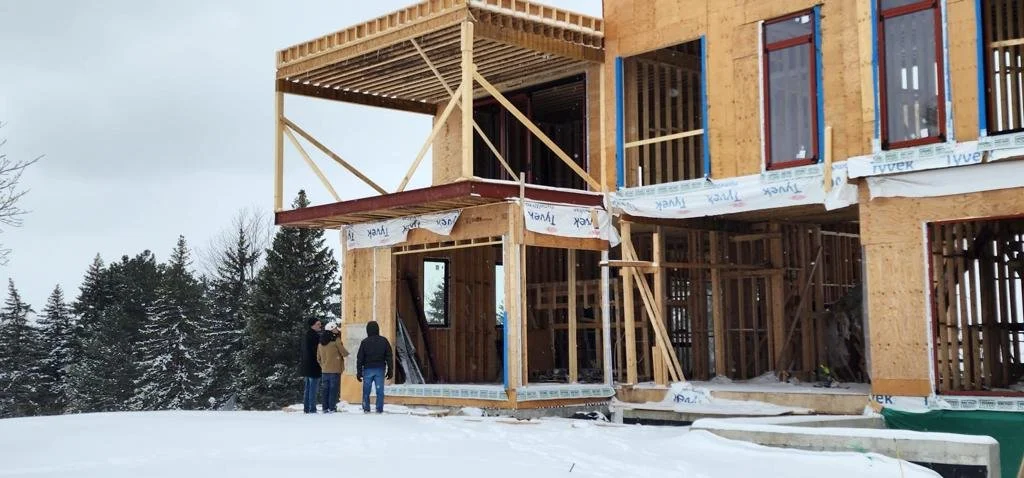Three people are standing outside a partially constructed wooden building in a snowy landscape with trees in the background.