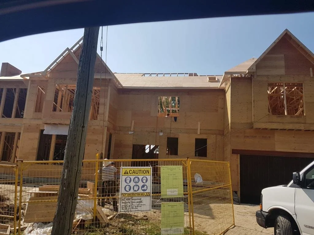 Under construction two-story wooden house with framing and partial roof, fenced off in front.