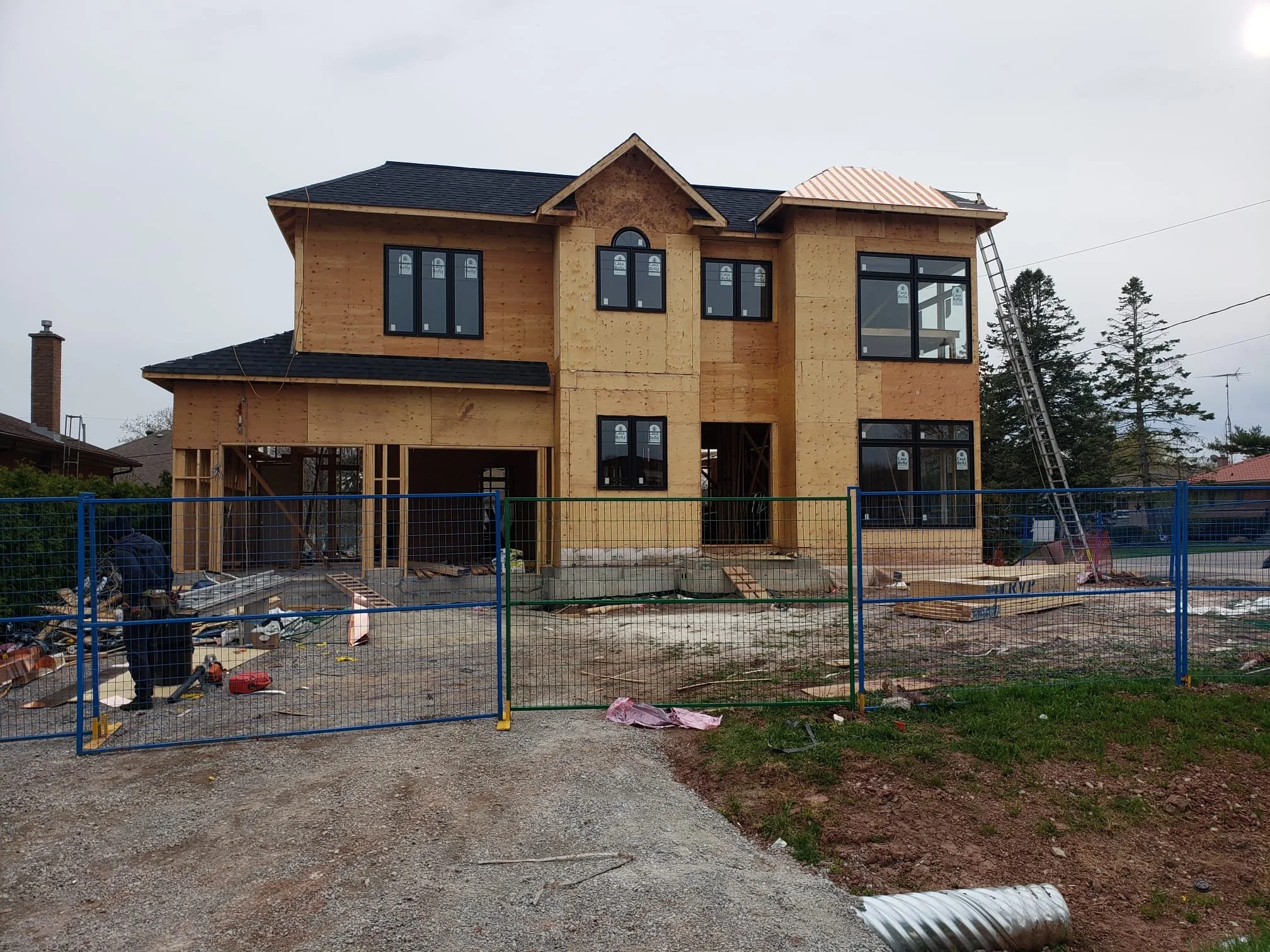 Two-story house under construction with wooden framing, black window frames, and a ladder leaning against the right side. Construction materials and equipment are scattered around the unfinished yard, enclosed by a blue and green fence.