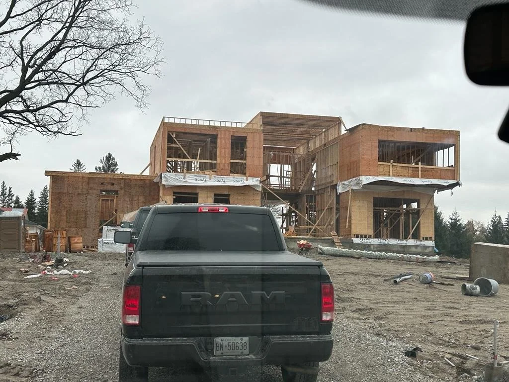 A two-story house under construction with a black RAM truck parked in front, surrounded by construction materials and debris on a gravel lot under an overcast sky.