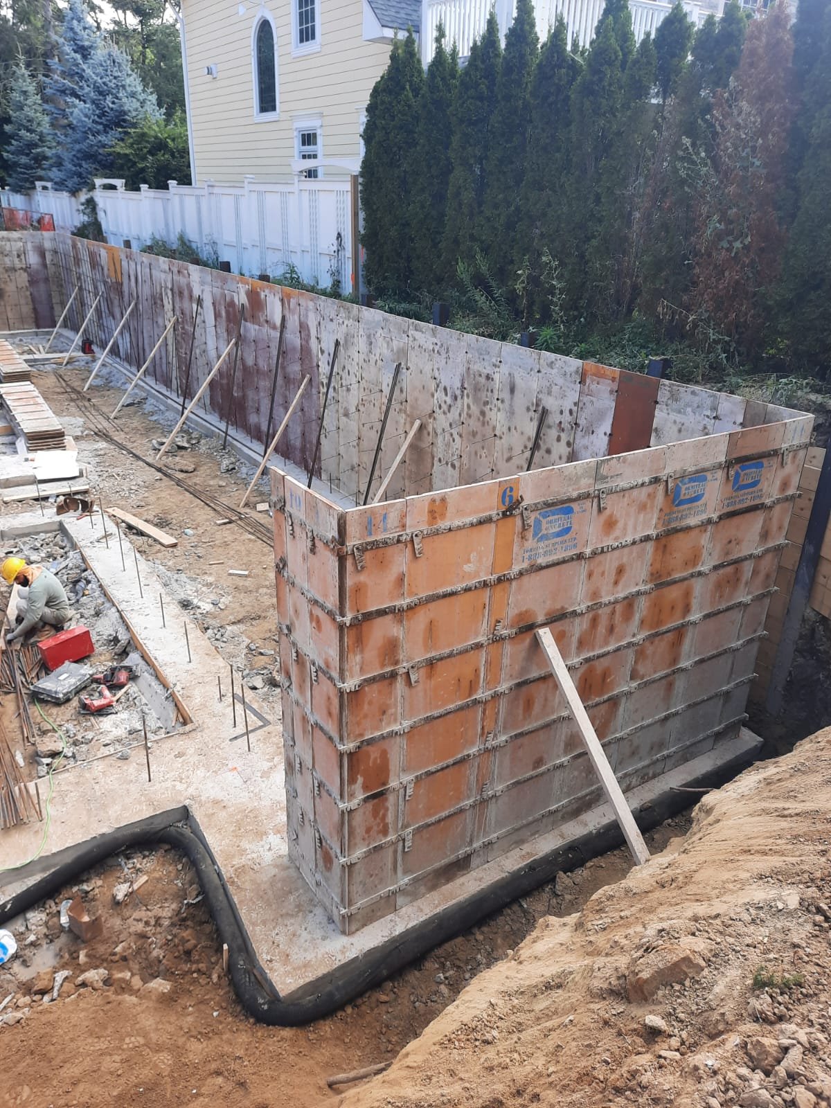 Construction site with brick wall forming foundation for a building, with worker wearing a yellow helmet working nearby.