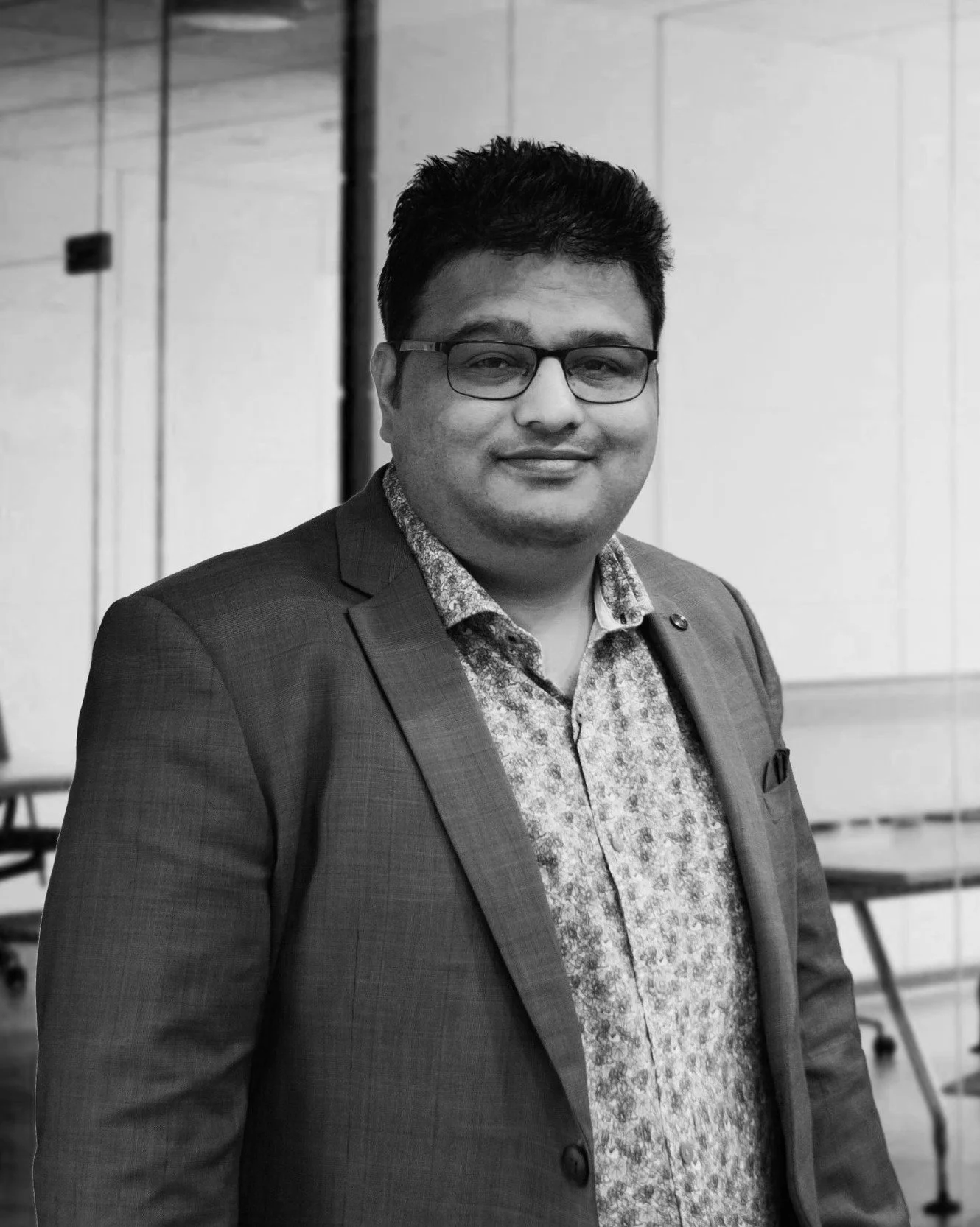A man wearing glasses, a patterned shirt, and a suit jacket standing indoors in front of chairs and a white wall.
