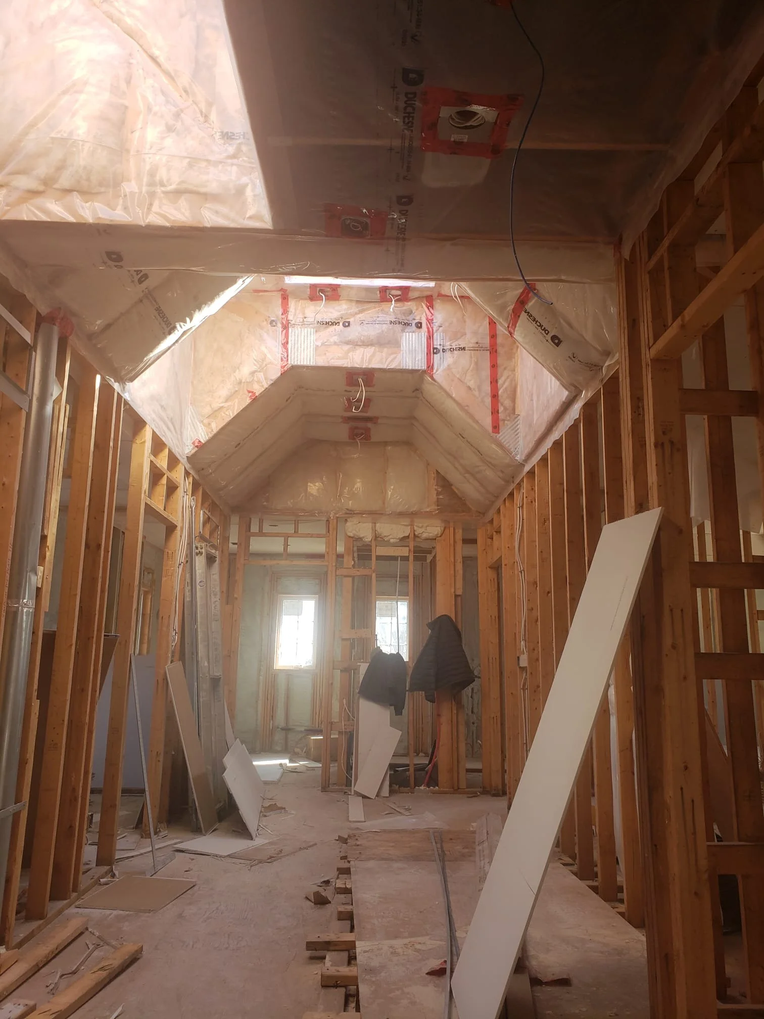 View inside a house under construction, showing exposed wooden framing, drywall sheets leaning against the walls, and a partially finished ceiling with insulation and venting.