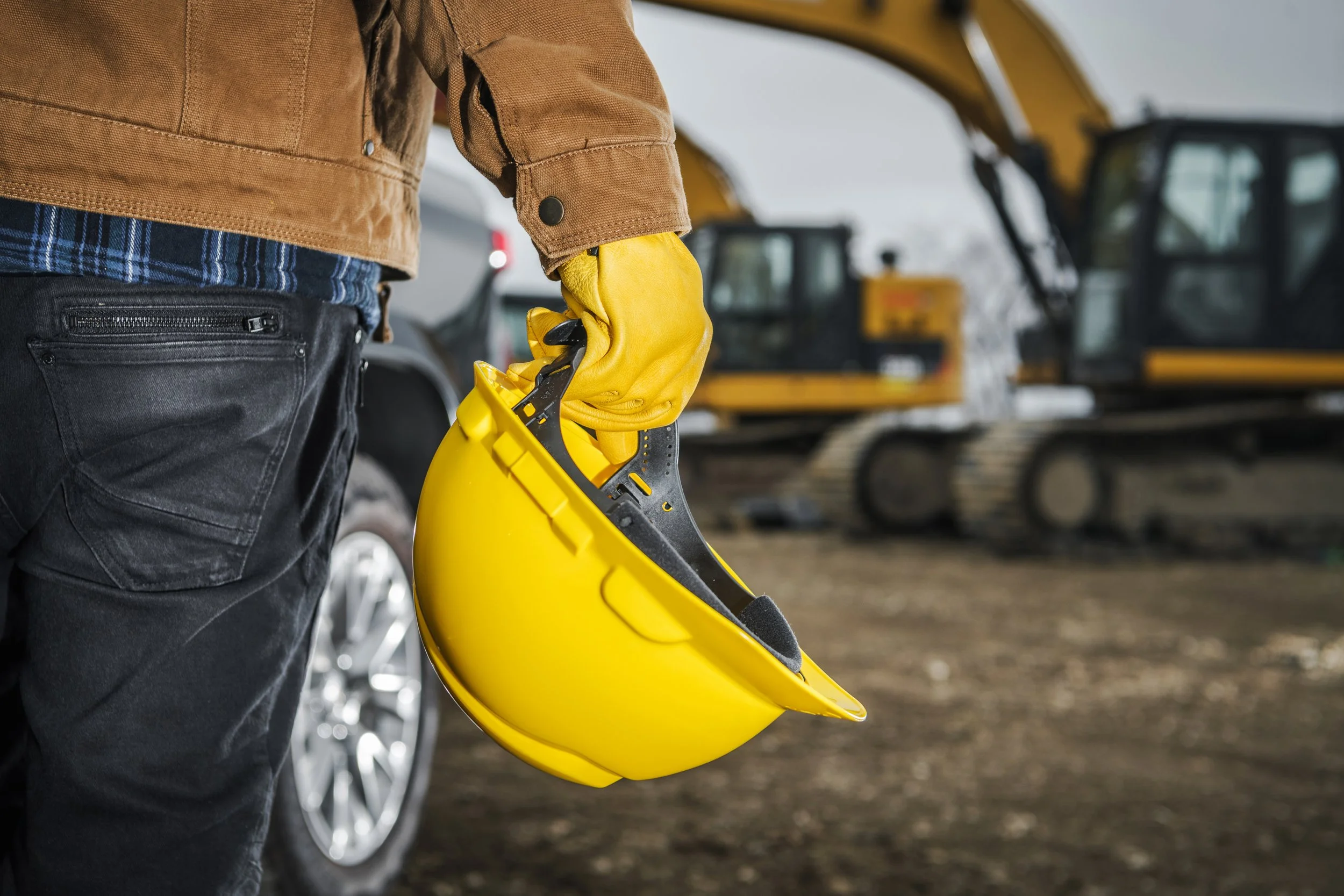 Person wearing a brown jacket, black jeans, and yellow gloves holding a yellow construction helmet with a black suspension system, standing on a construction site with yellow excavators in the background.