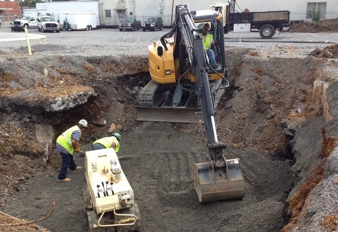 Construction workers and a yellow excavator working on a large excavation site, pouring concrete or filling with gravel.