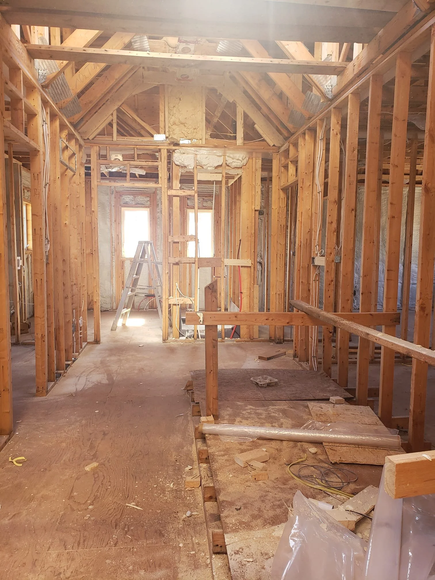 Interior of a house under construction with exposed wooden framing, a ladder, and construction materials scattered across the unfinished floor.