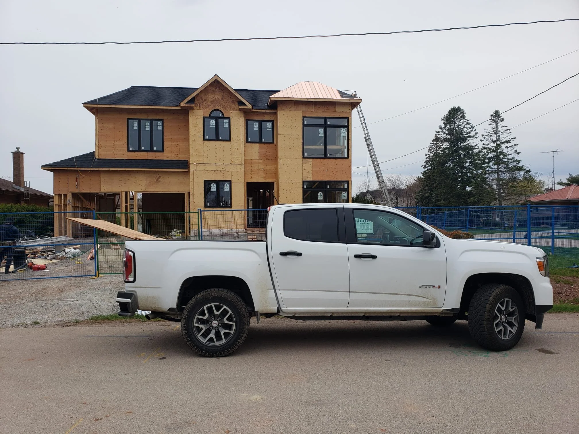 A white pickup truck parked in front of a house under construction, with part of the house's wooden frame and exterior walls visible, and a ladder leaning against the house.