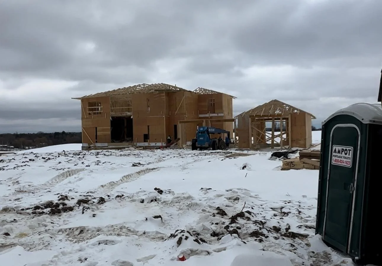 Under construction two-story wooden house in a snowy landscape, with a portable toilet on the right.