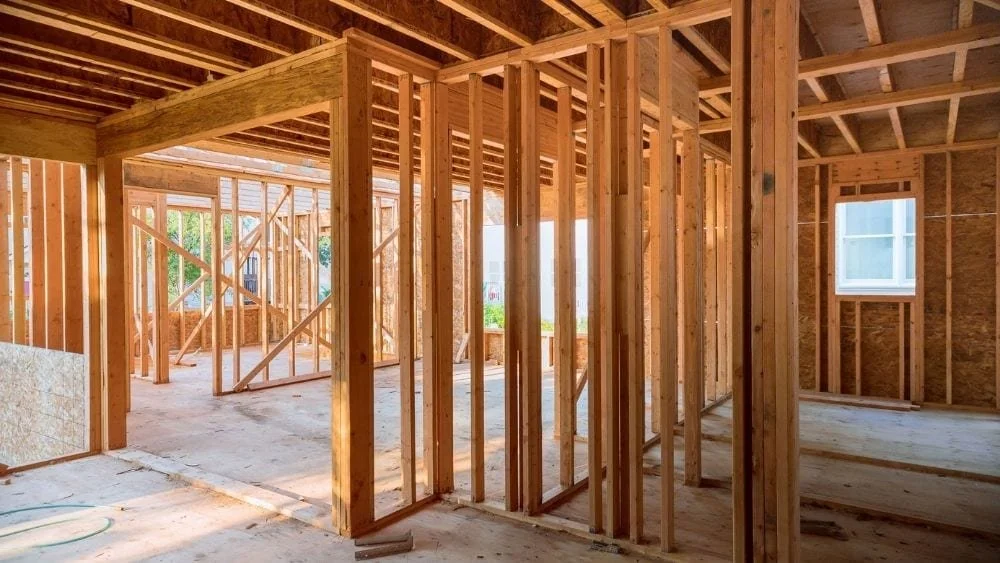 Interior view of a house under construction with exposed wooden framing, studs, and ceiling beams, and a window on the right wall.