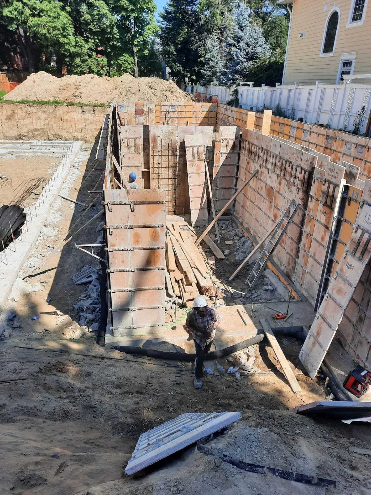 Construction worker walking on a dirt site with brick walls being built around a foundation, and construction materials scattered around.