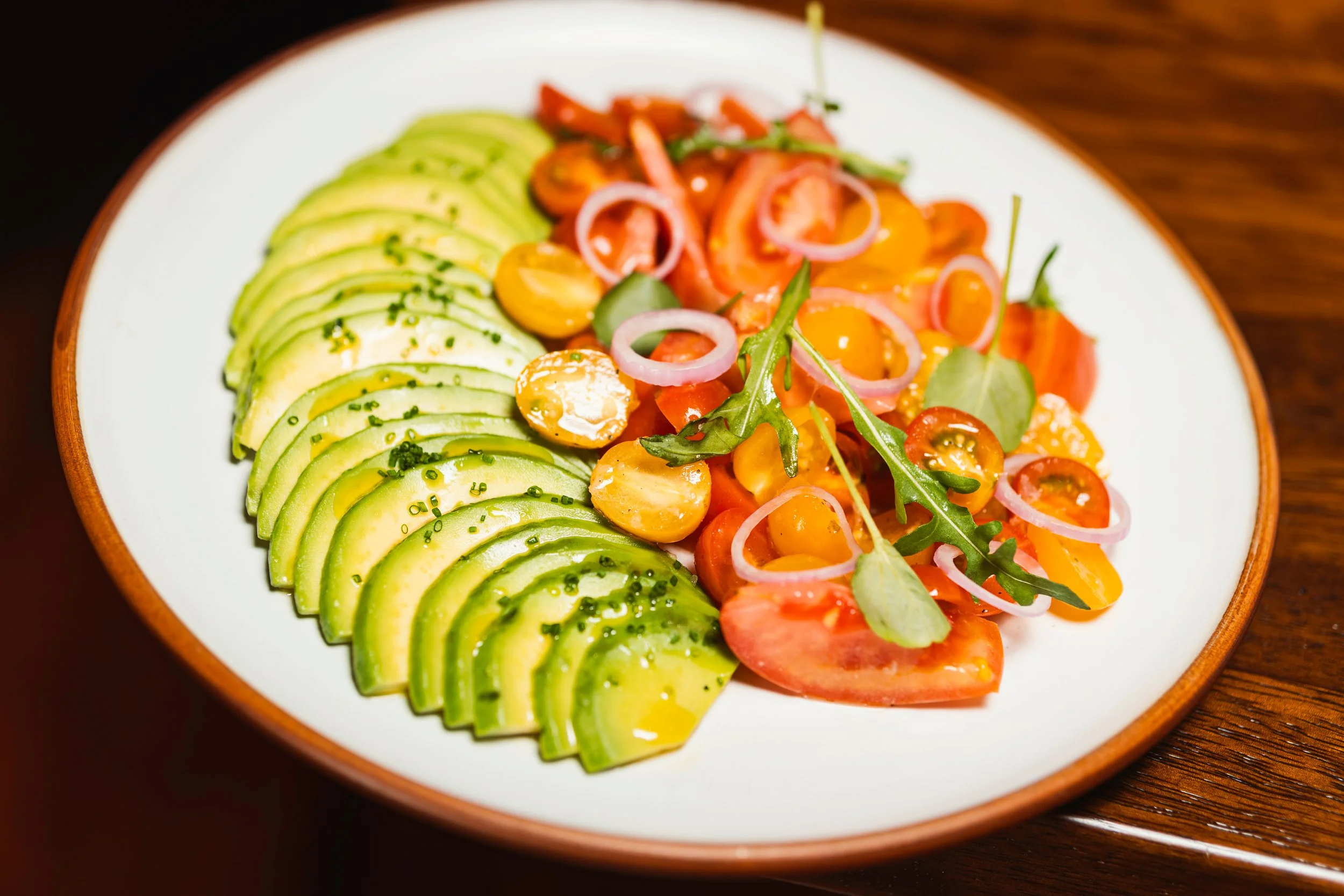 En un plato de cerámica blanca, en un lado hay rebanadas de aguacate con perejil picado, y en el otro lado hay ensalada con tomates cherry, hojas de arúgula y cebolla en rodajas, todo preparado para comer.