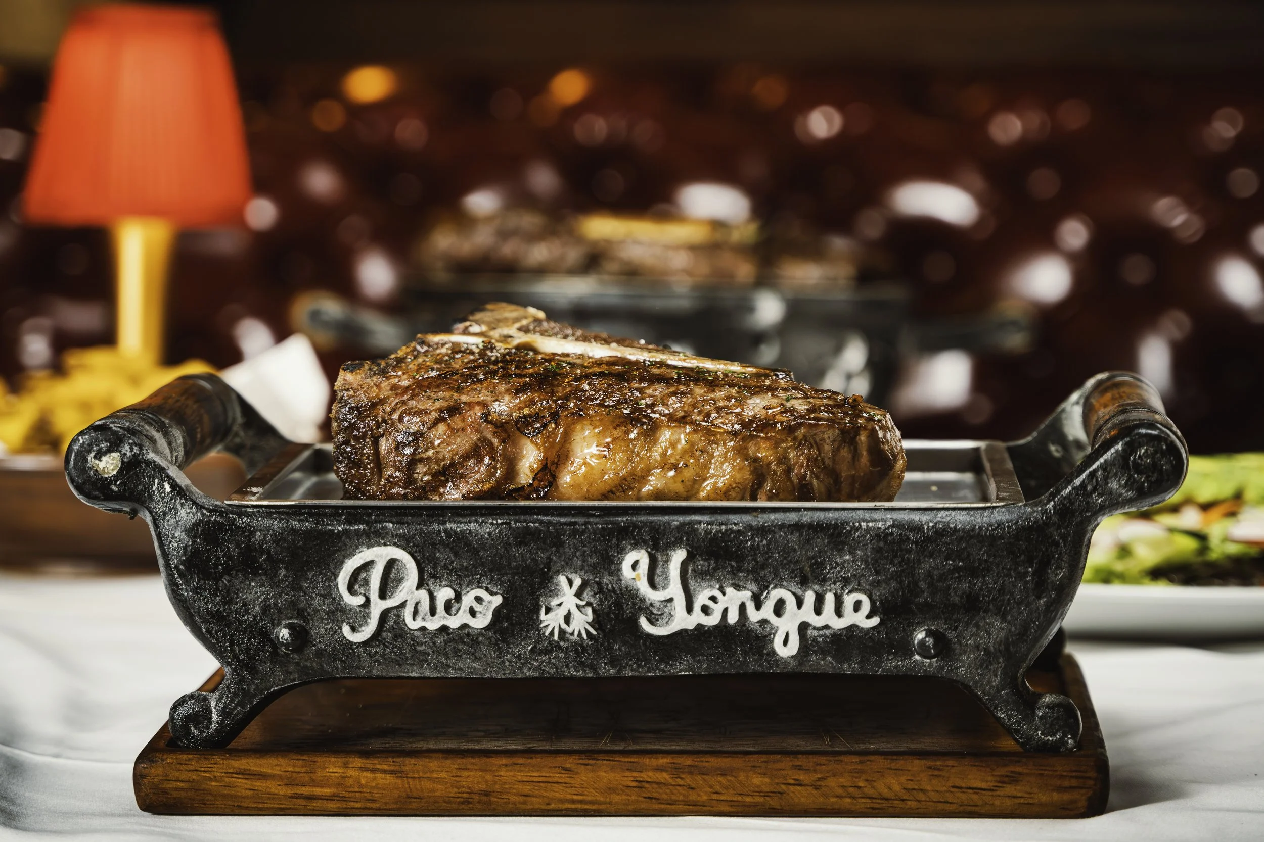 Un corte de carne asada en una bandeja de hierro sobre una base de madera, con un fondo borroso y una lámpara roja a la izquierda.