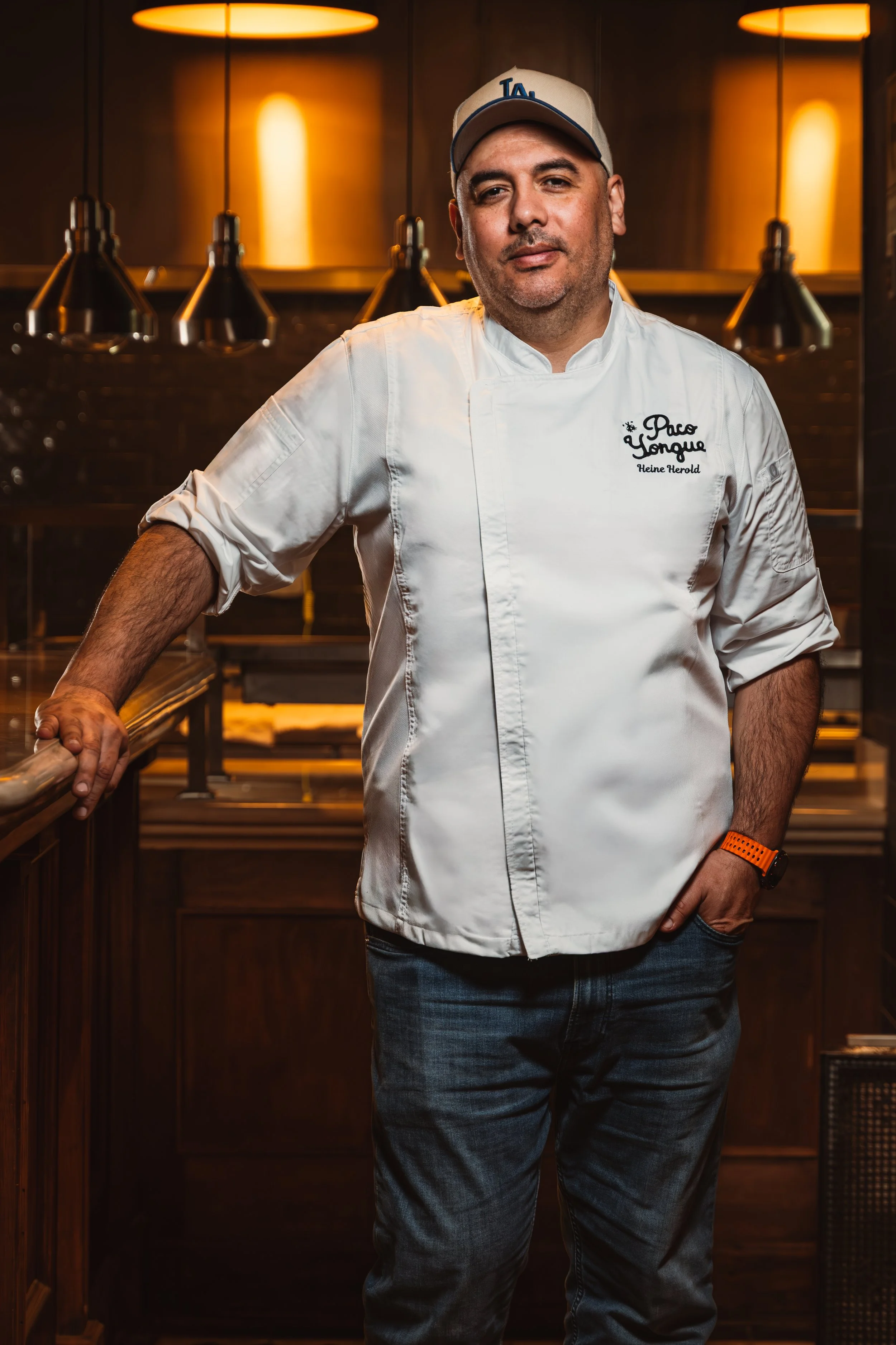 Hombre con uniforme de chef, con gorra y camisa blanca, en una cocina o restaurante, con iluminación cálida.