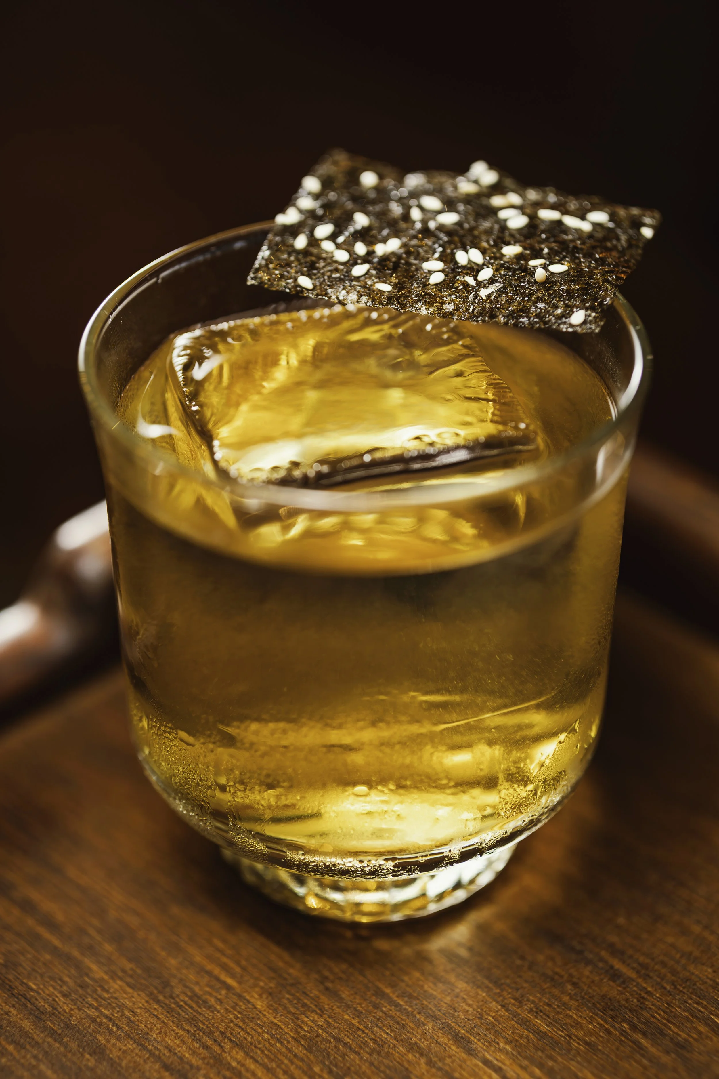 Vaso con bebida dorada, hielo y una galleta con semillas de sésamo en la orilla del vaso.