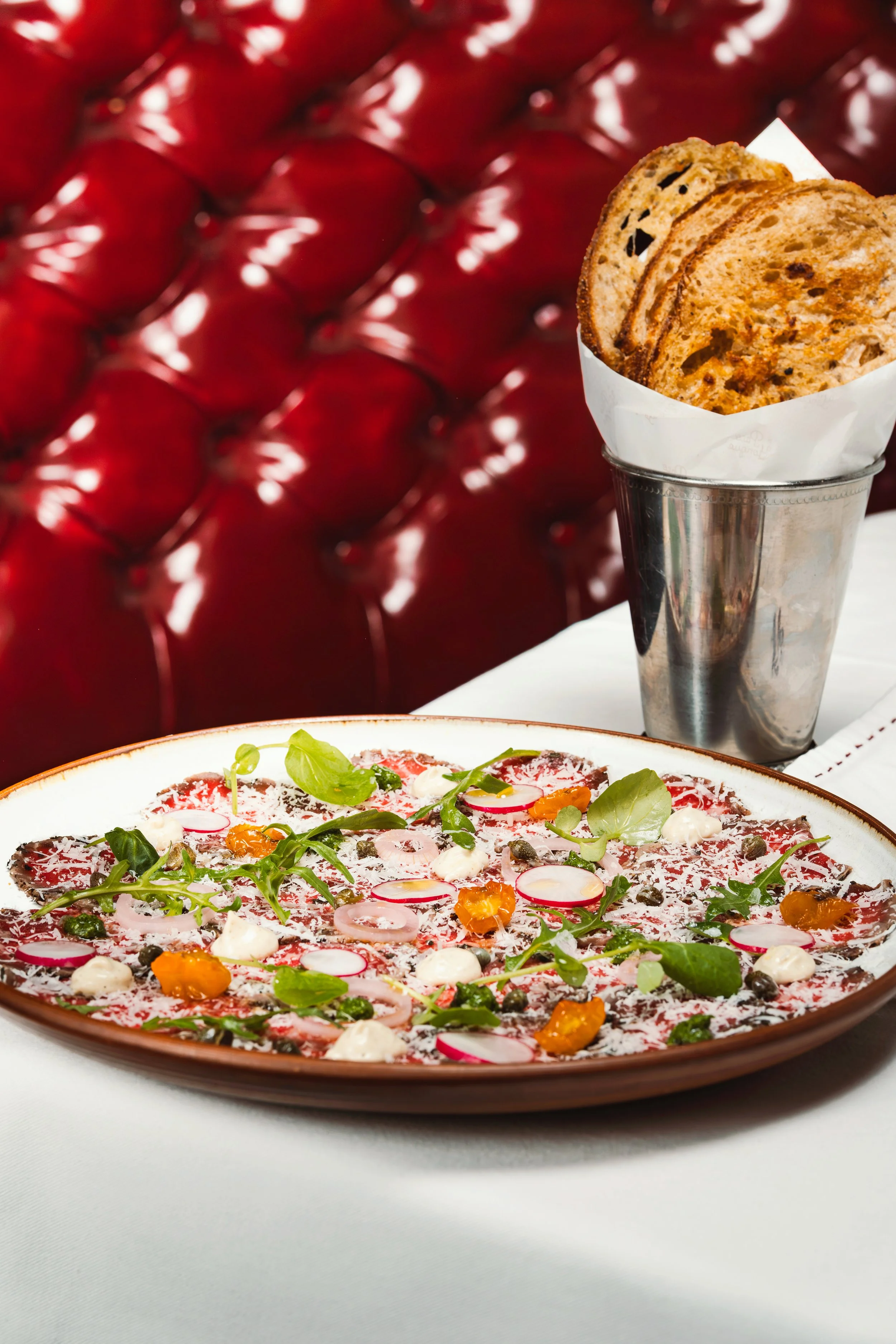 Una pizza fina y decorada con rodajas de rábano, hojas verdes, crema y semillas, acompañada de pan tostado en un vaso metálico, en un fondo de tapiz de cuero rojo.