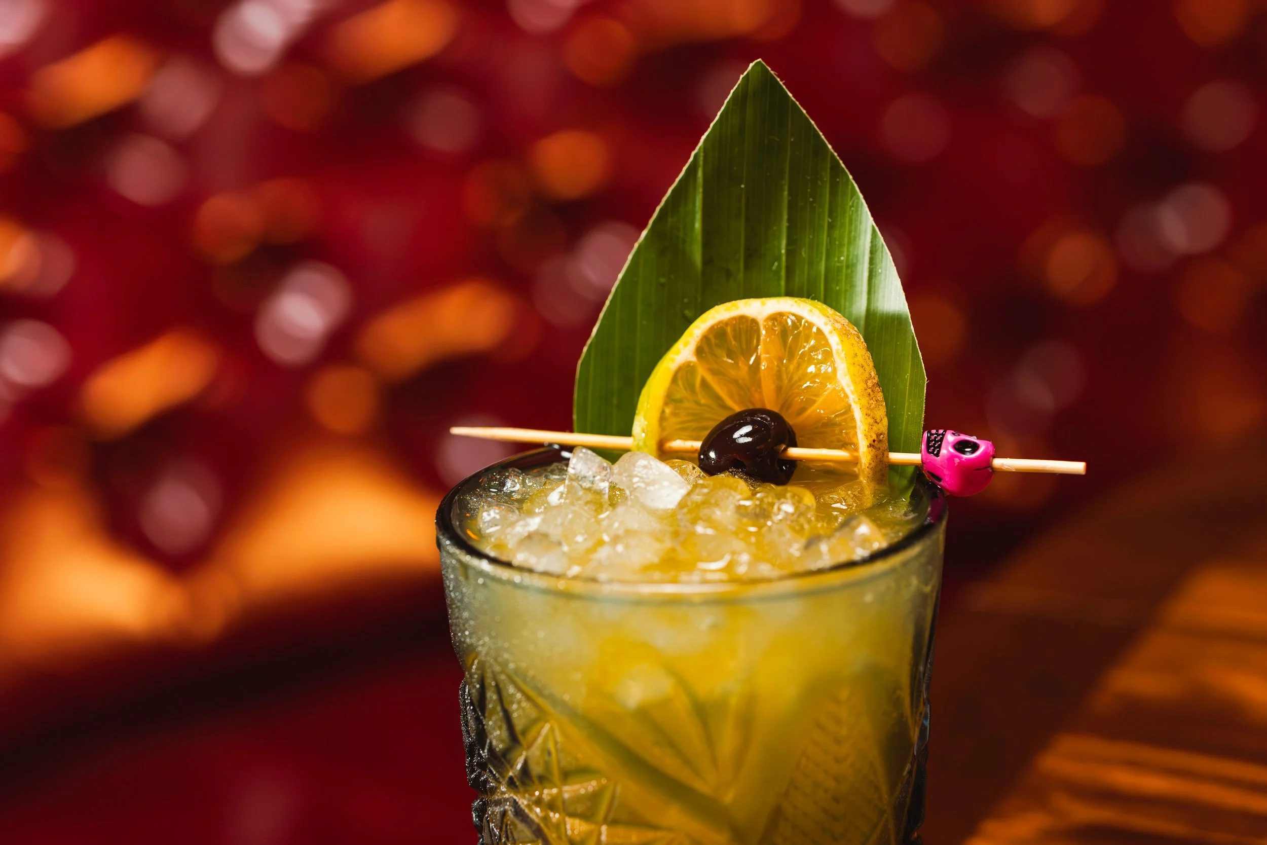 Cerveza fría con rodaja de limón, una aceituna y decoración de hoja verde en un vaso cristalino con hielo, en un fondo difuso y cálido.