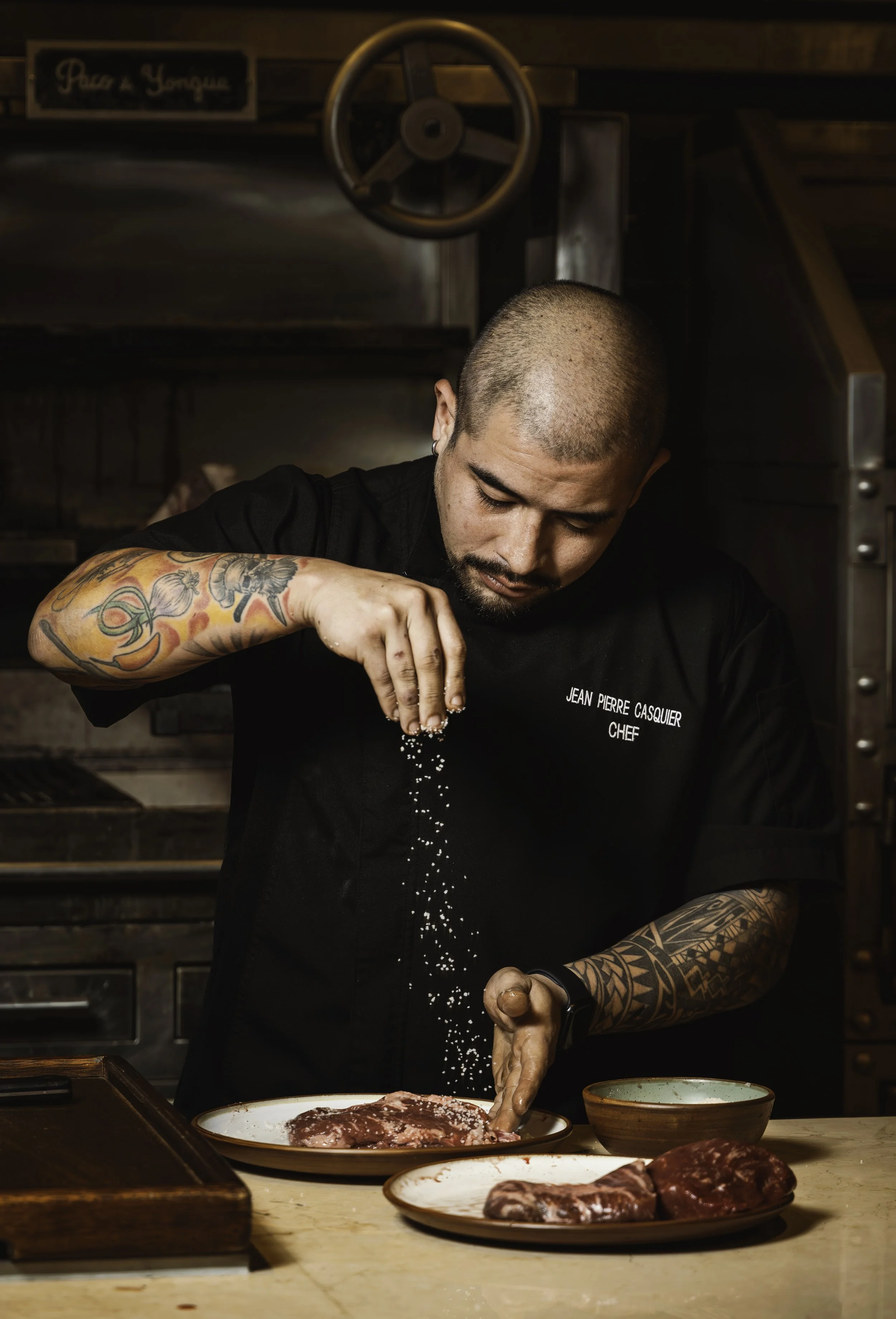 Un chef con tatuajes sazonando carne en una cocina rústica.