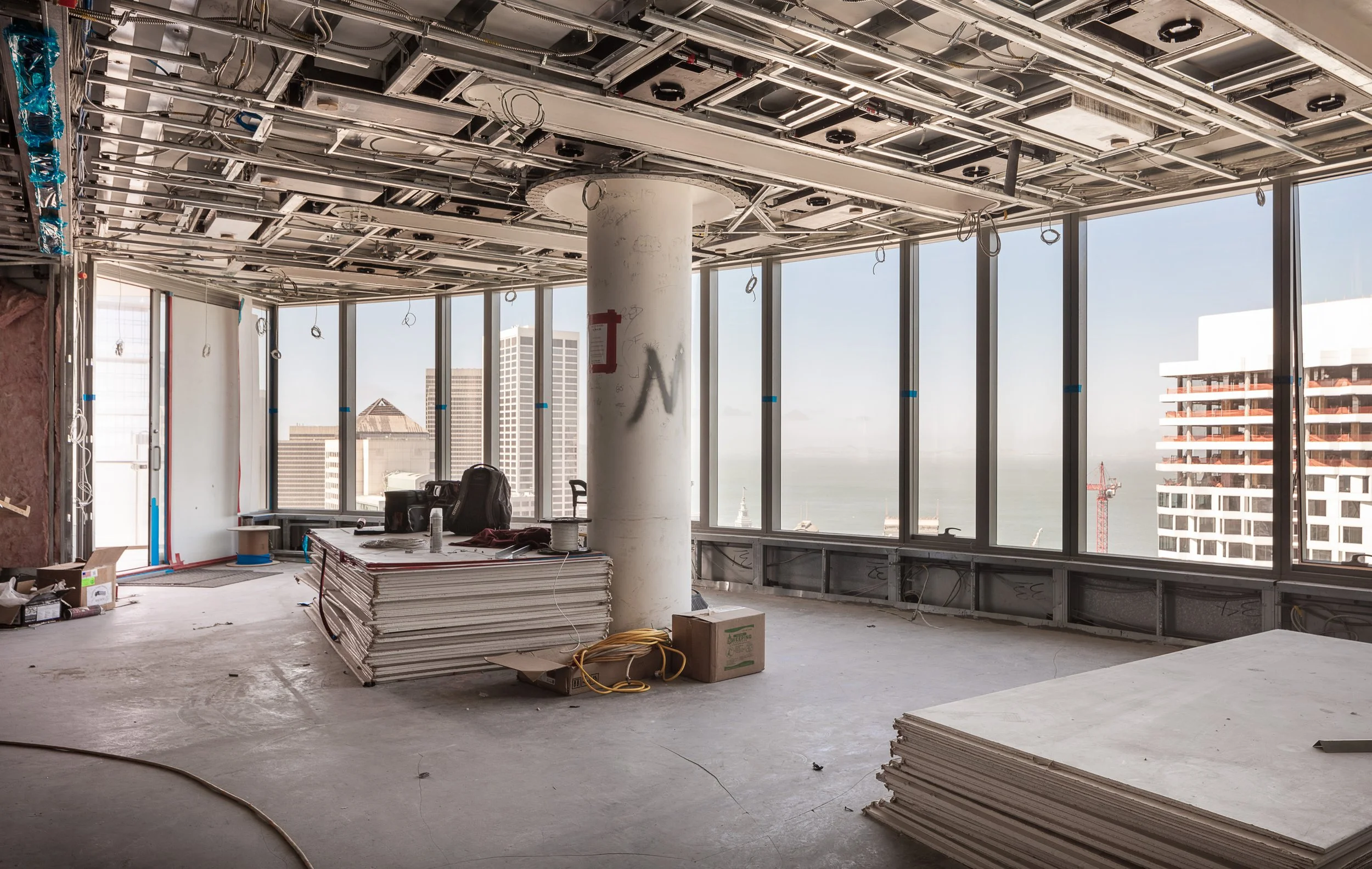 Interior of a high-rise building under construction with large windows showing a city skyline and water in the distance, exposed ceiling framework, construction materials, and tools scattered on the floor.