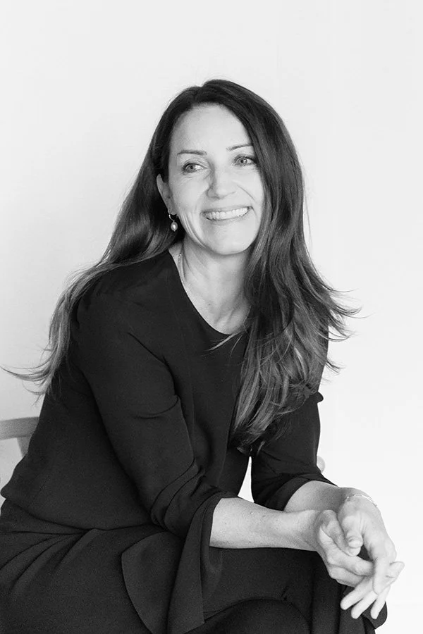 Black and white photo of a smiling woman with long hair, wearing a dark dress, sitting with her hands clasped, against a plain background.