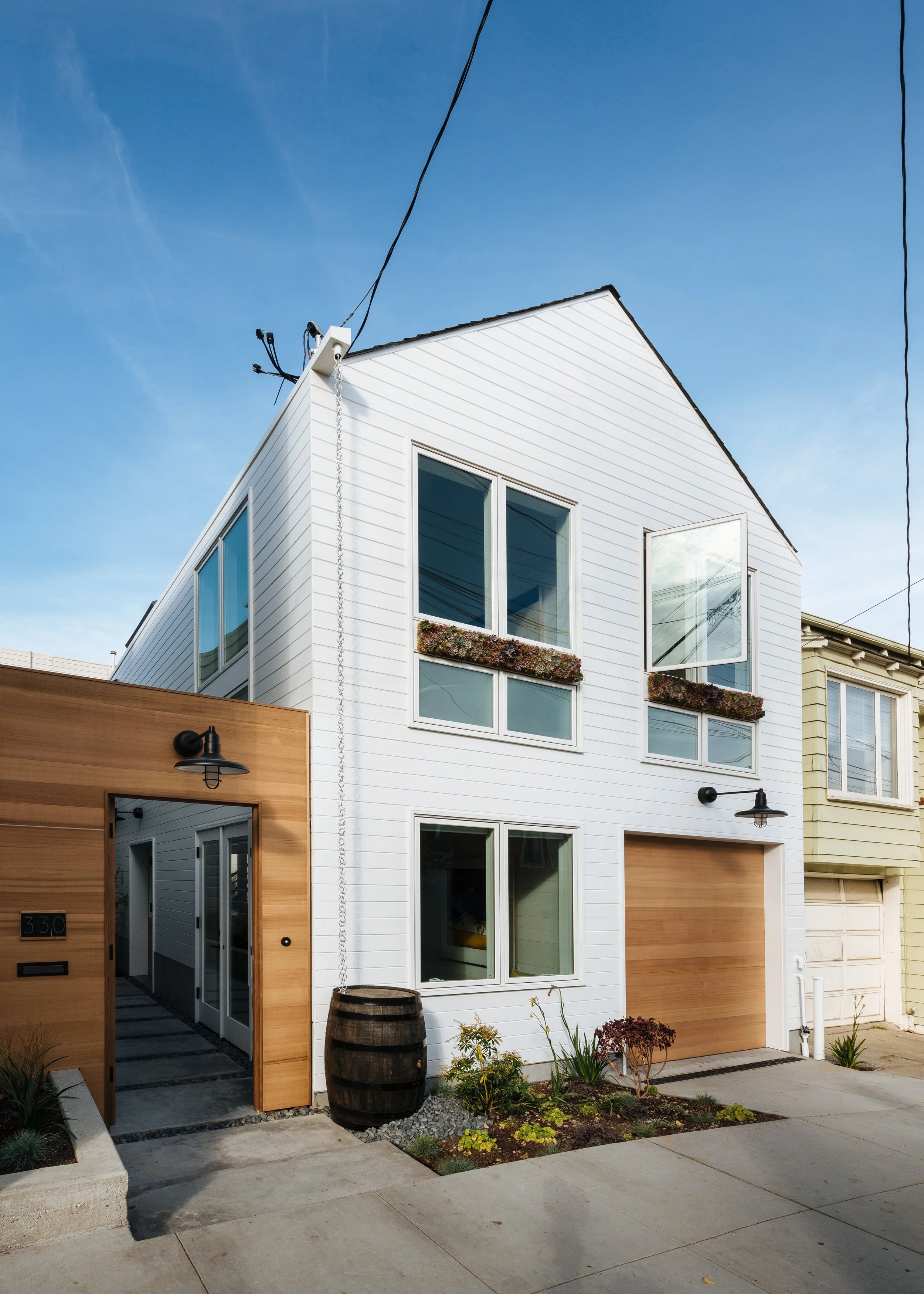 A modern white multi-story house with large windows, a wooden garage door, and a small garden area in front, under a blue sky.