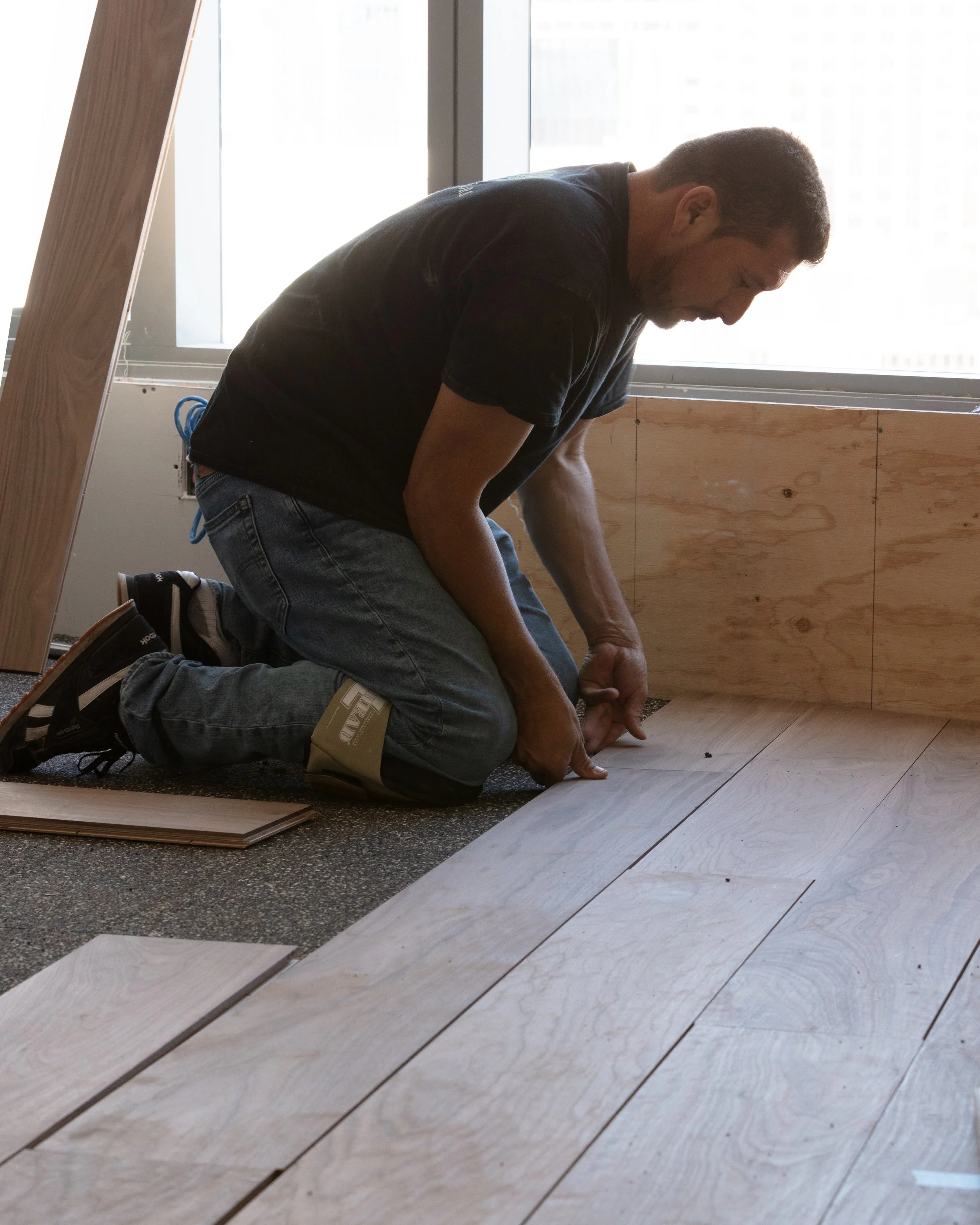 A man kneeling on the floor installing wooden floorboards near a large window, with additional flooring materials nearby.
