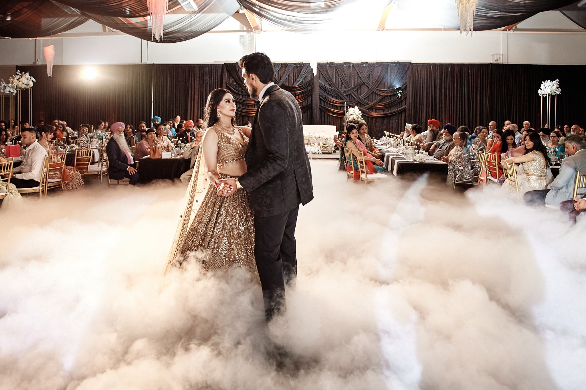 A bride and groom are dancing in the center of a wedding reception with a fog effect on the floor. Guests are seated at tables in the background under a decorated ceiling.