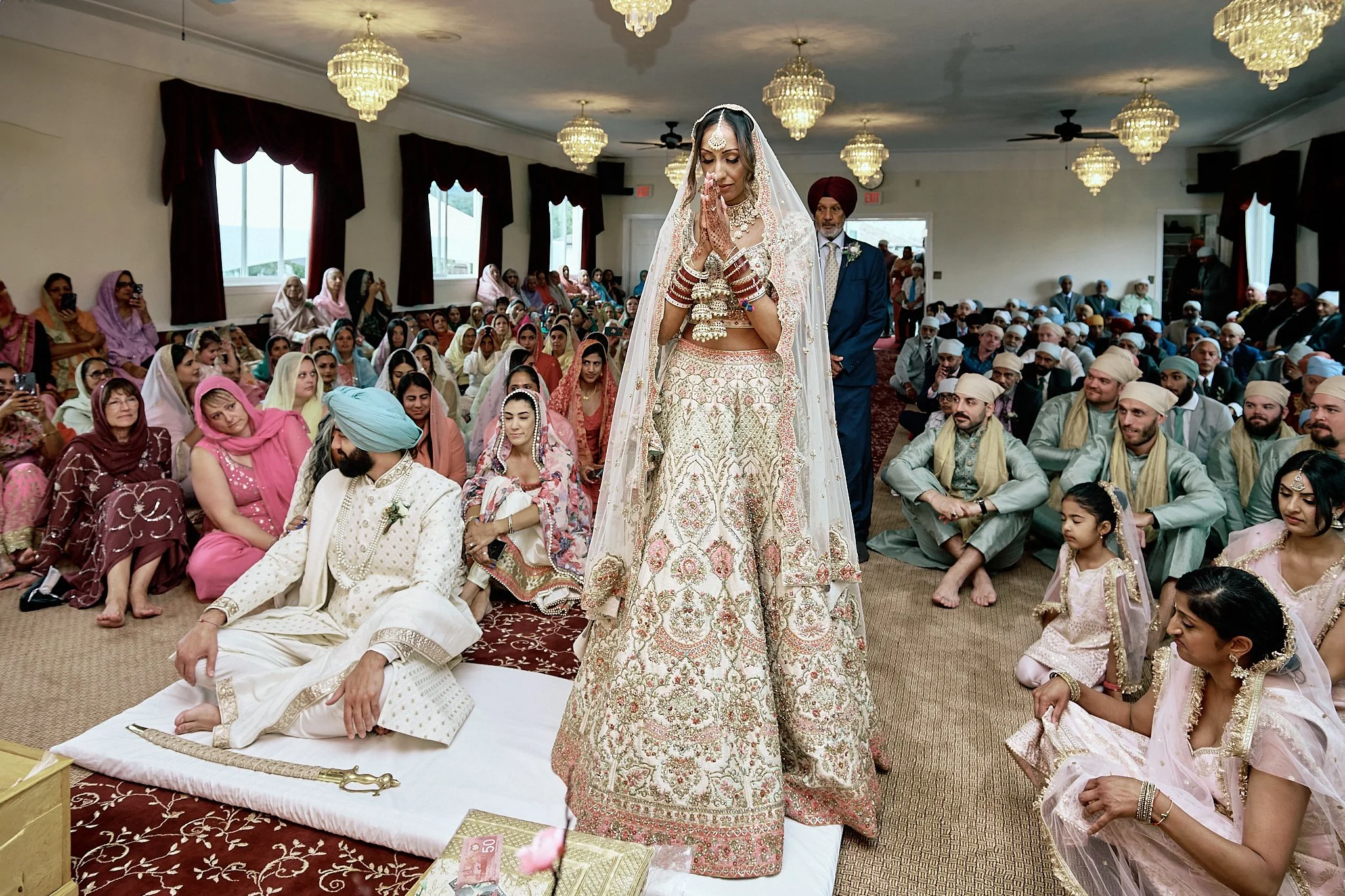 A bride and groom participating in a traditional Sikh wedding ceremony indoors, with guests seated around them. The bride is wearing a heavily embroidered bridal lehenga, jewelry, and a veil, standing with hands in prayer. The groom, wearing a cream sherwani and a turban, sits on the floor with one hand resting on his knee. Guests are dressed in colorful traditional attire, some sitting on the floor, others in chairs, with some taking photos.