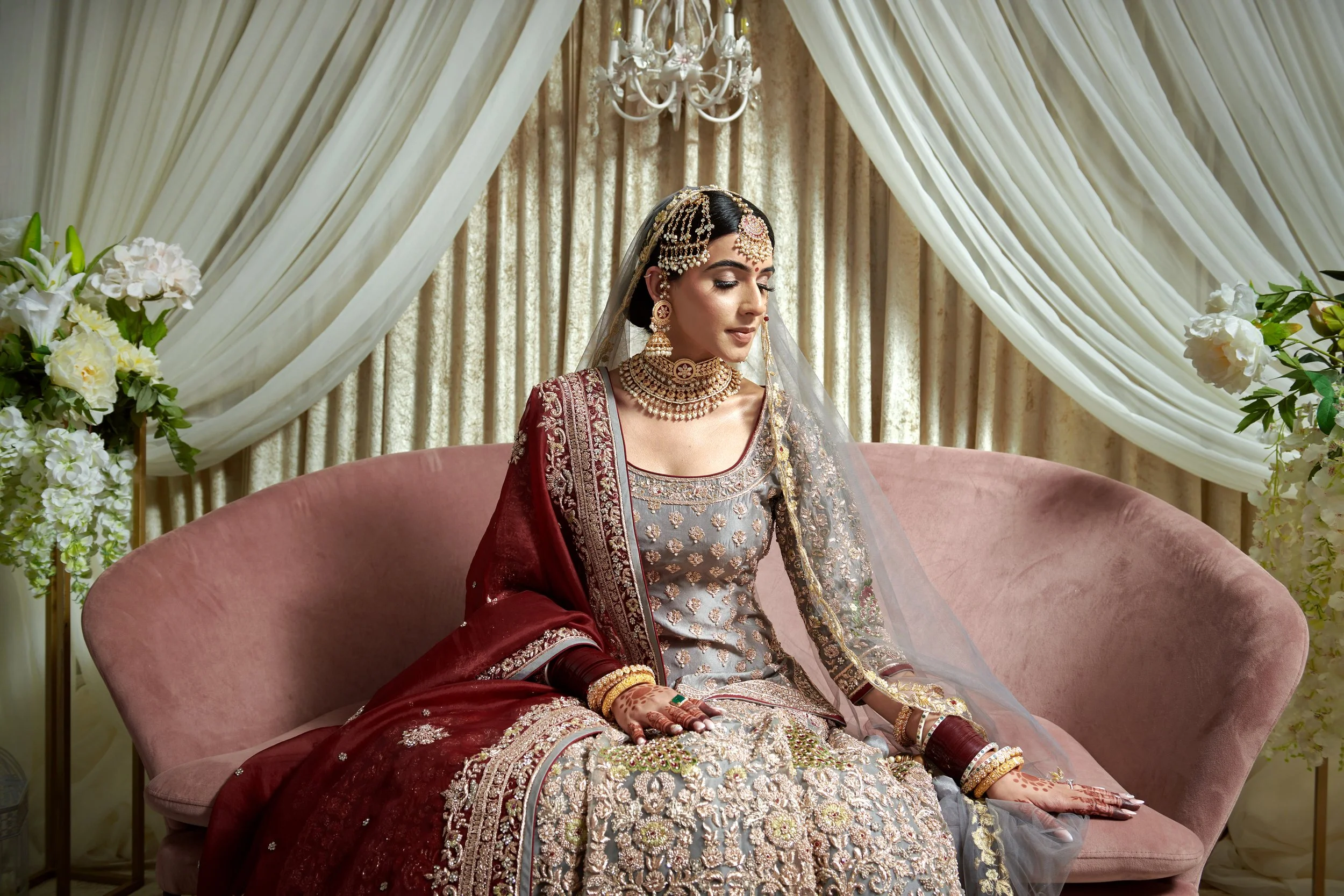 A woman dressed in traditional Indian bridal attire sitting on a pink sofa with cream-colored drapes and floral arrangements in the background.