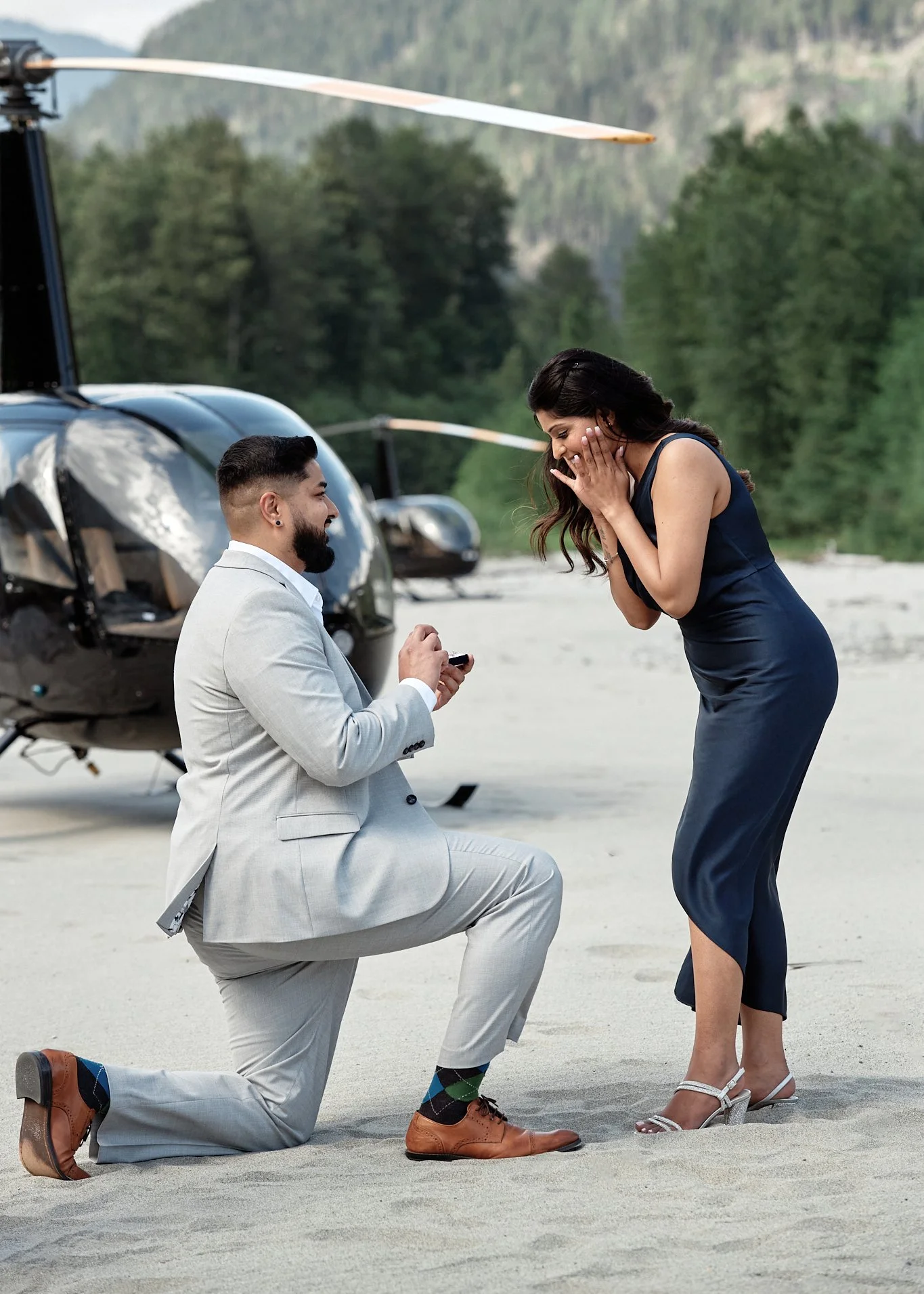 A man proposing marriage to a woman on a beach with a helicopter in the background, the woman looks surprised and happy.