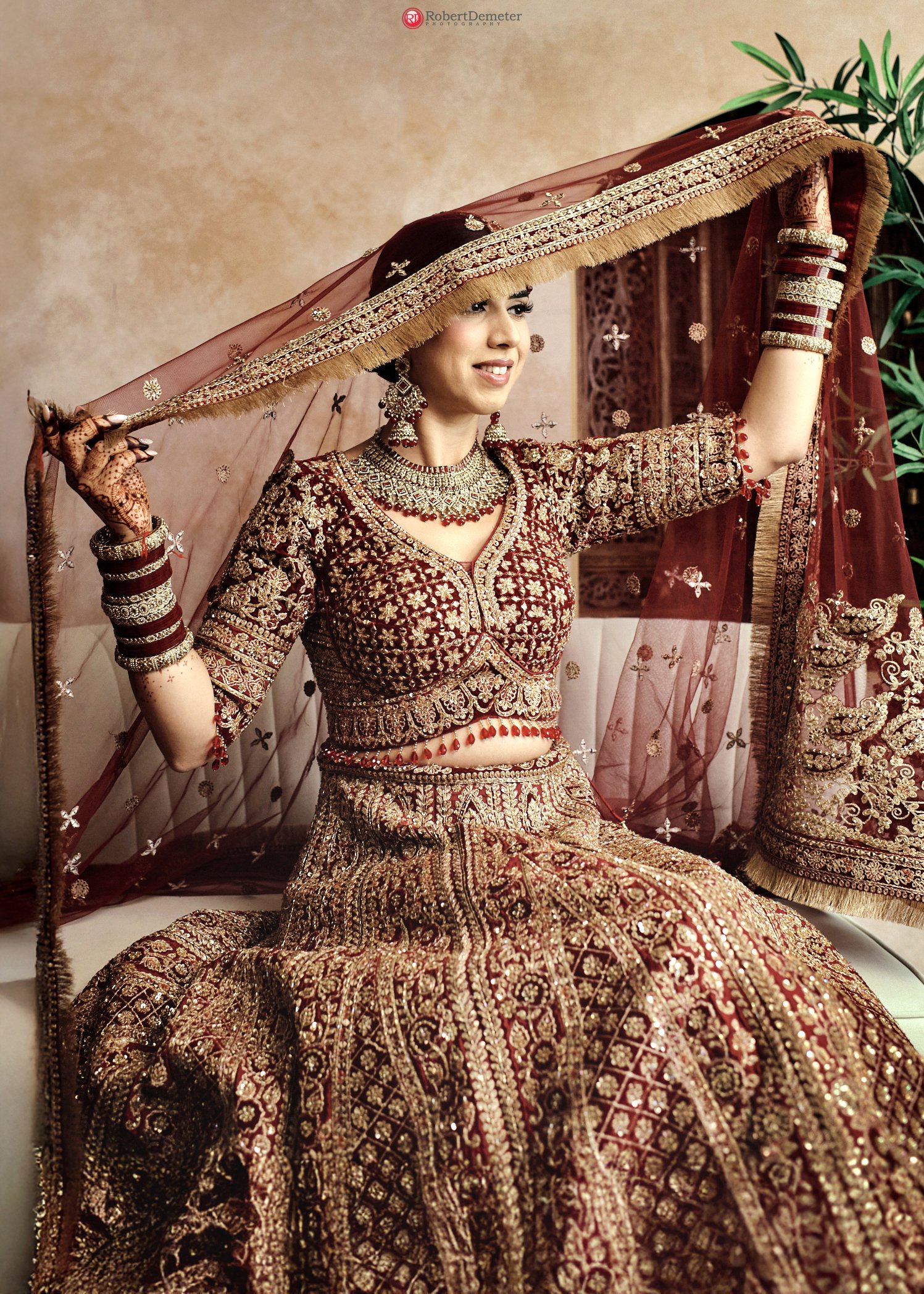A woman dressed in traditional Indian bridal attire, wearing a heavily embroidered maroon and gold lehenga, jewelry, and a large veil, sitting on a sofa with a beige wall and green plant in the background.