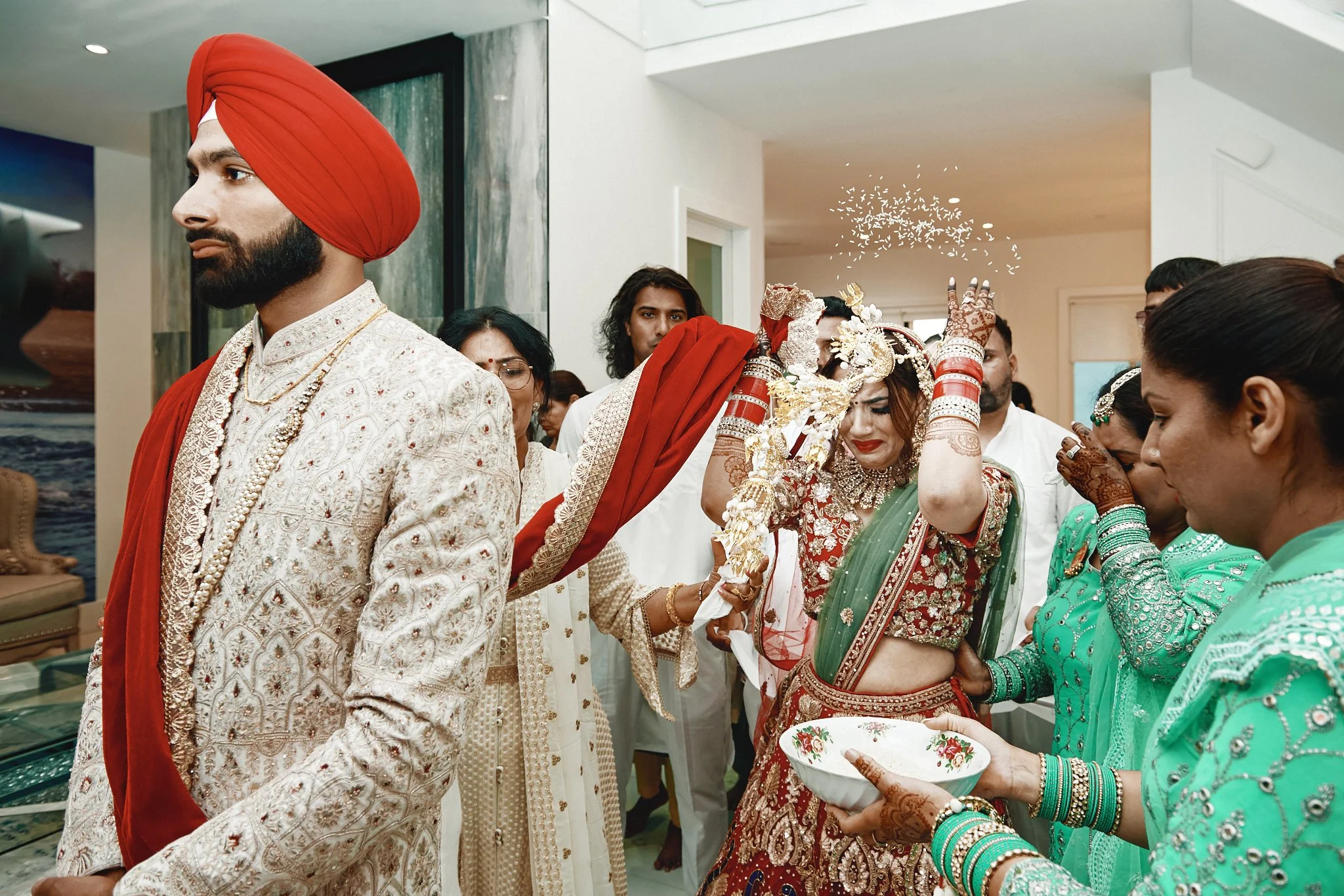 Indian wedding ceremony with bride in traditional red and gold attire, surrounded by women in turquoise dresses, during a ritual with rice thrown in the air.