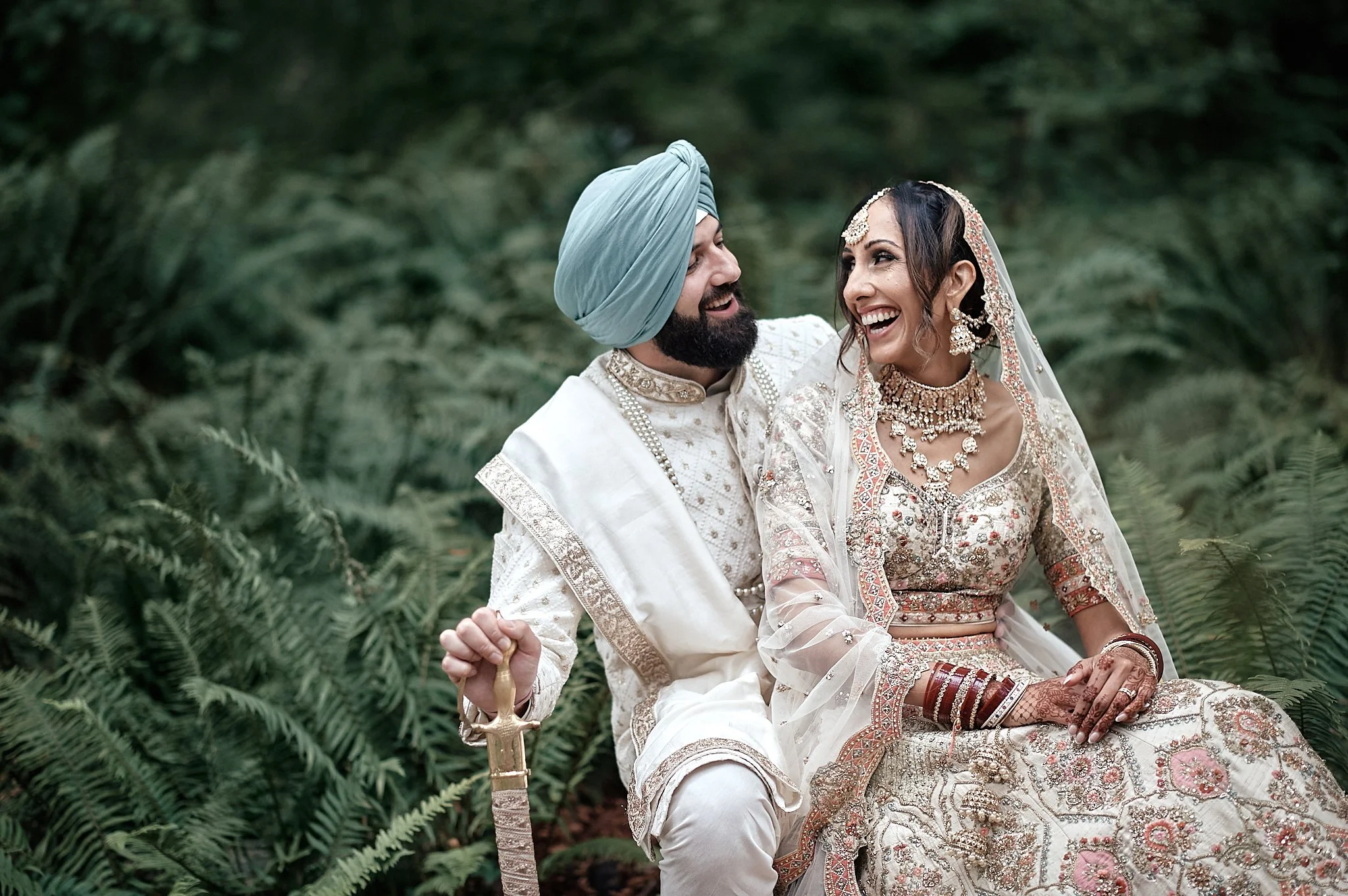 A newlywed Indian couple in wedding attire sitting outdoors on green foliage, smiling and looking at each other.