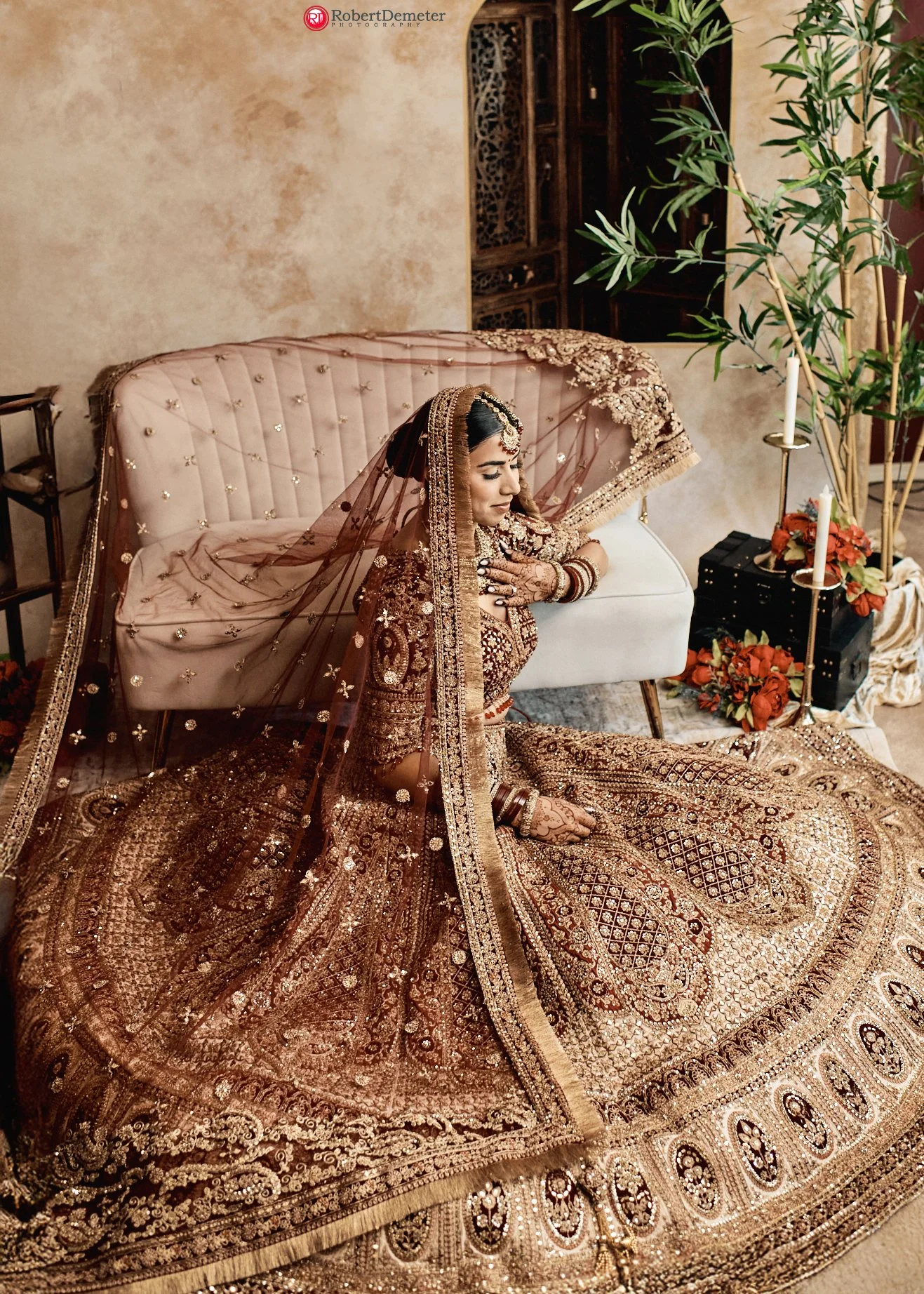 A bride dressed in a heavily embroidered, traditional wedding outfit, sitting on the floor with her eyes closed, wearing jewelry and a veil, in a decorated indoor setting.