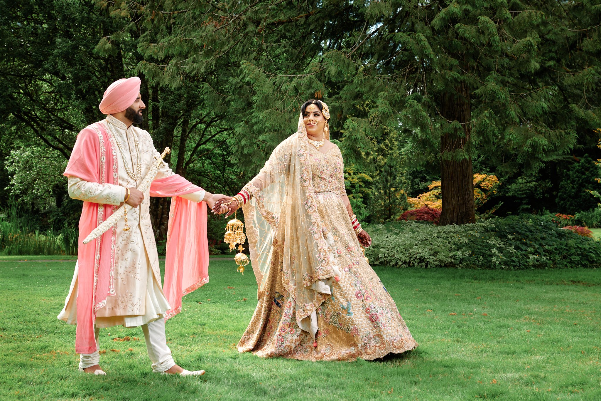 A bride and groom in traditional Indian attire holding hands in a lush green garden.