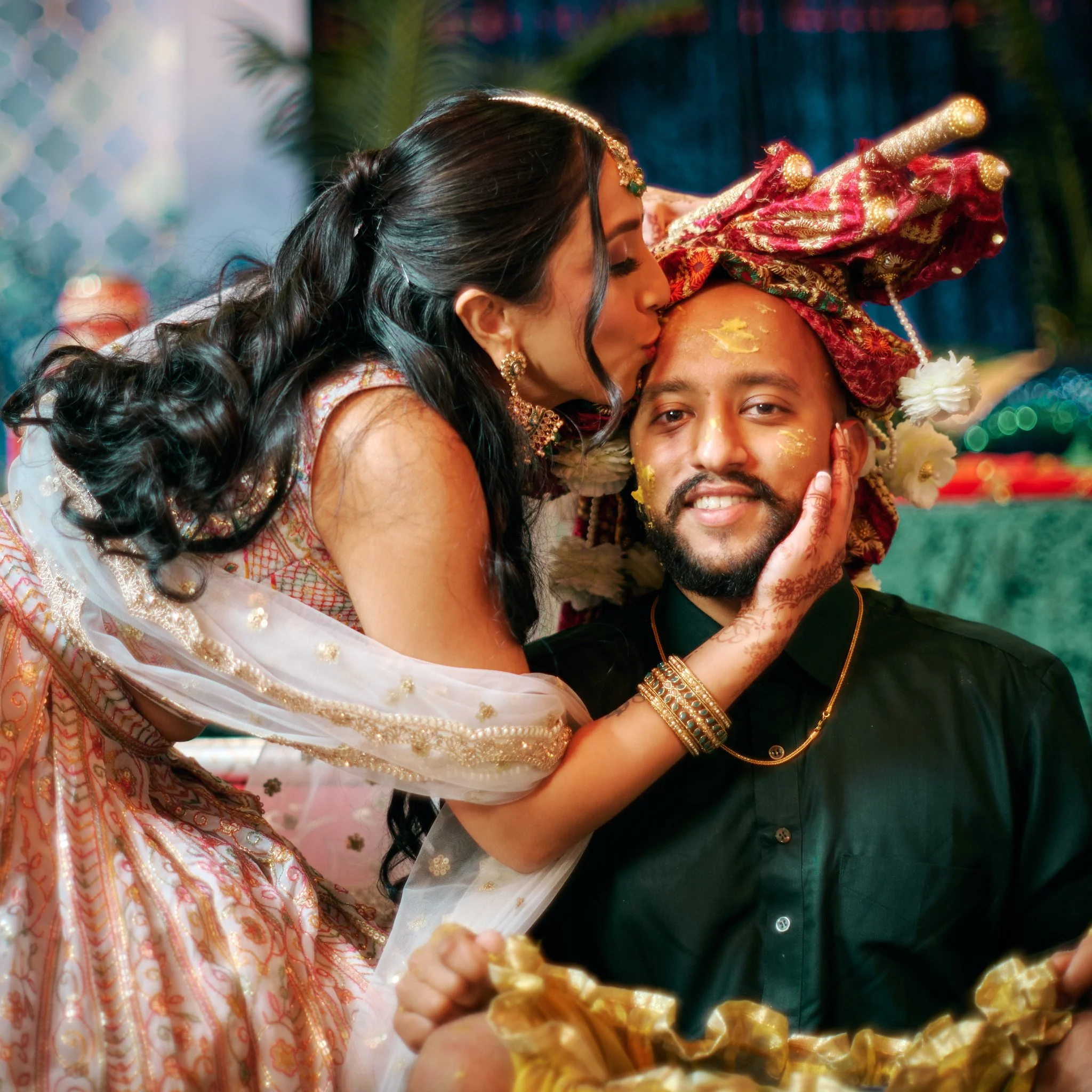 A woman in traditional Indian attire kisses a man on the forehead during a wedding ceremony, with the man wearing a colorful turban and gold jewelry.
