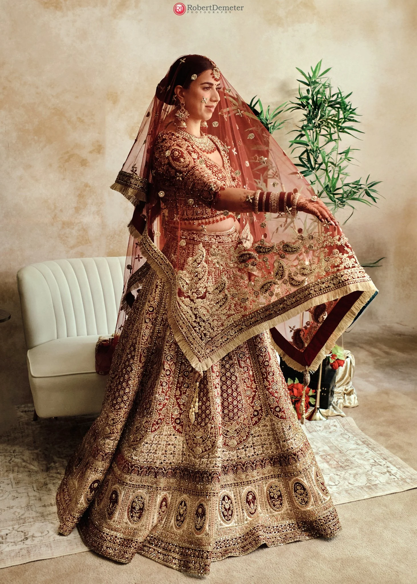 A woman in traditional Indian wedding attire, including a heavily embroidered maroon and gold lehenga, jewelry, and a veil, is posing indoors with a plant and a white sofa in the background.