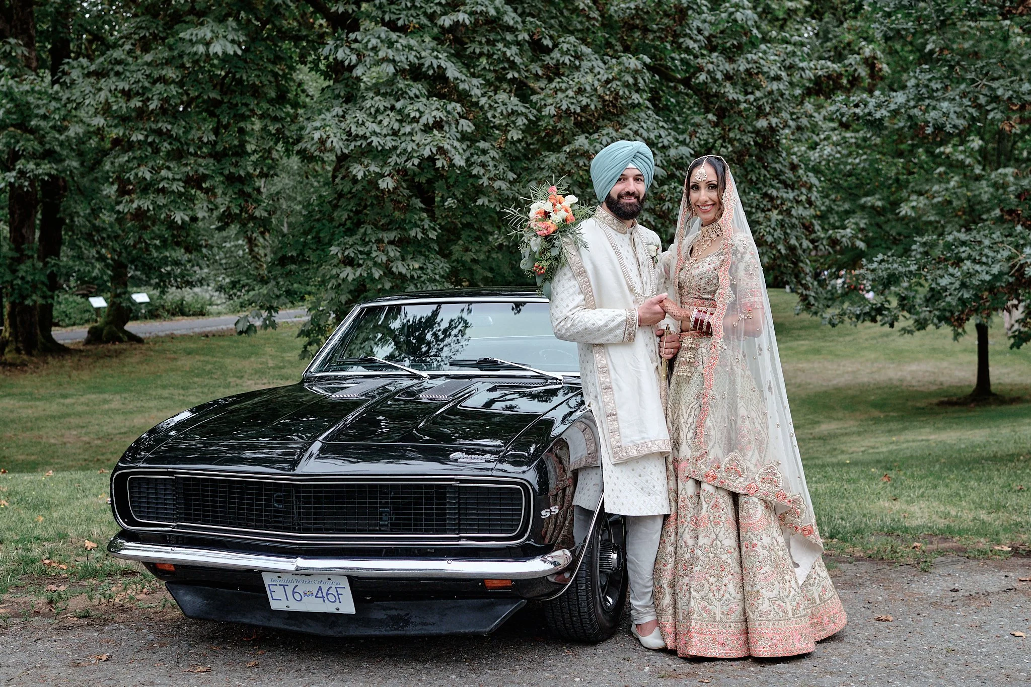 A newlywed South Asian couple dressed in traditional wedding attire standing outdoors next to a black vintage car with a background of green trees.