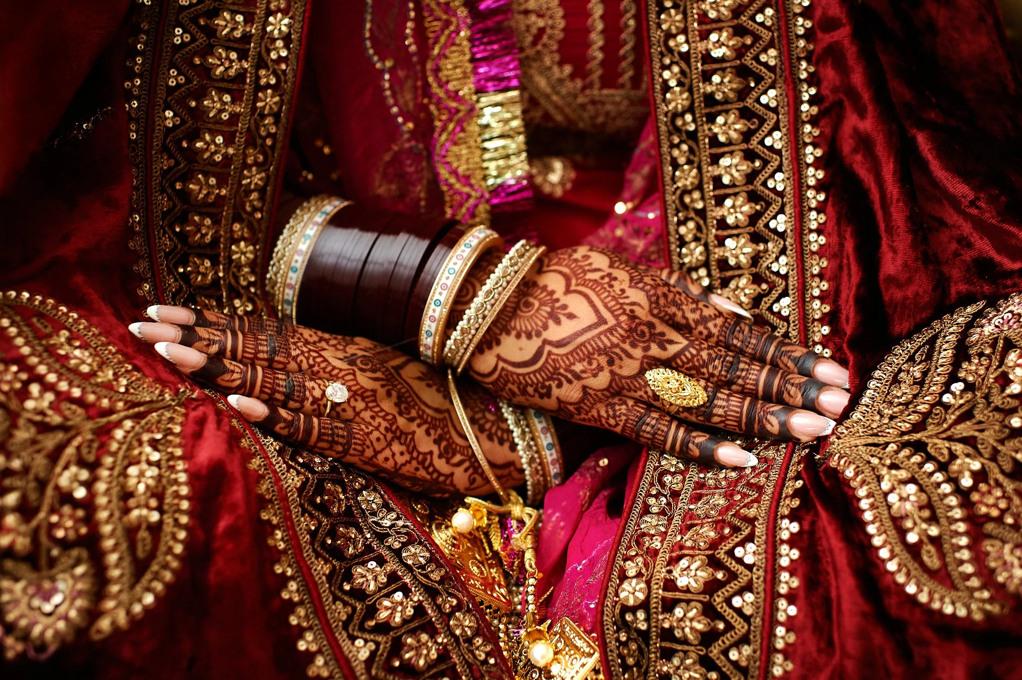 Close-up of a woman's hands resting on her lap, decorated with intricate mehndi (henna) designs, multiple bangles, rings, and jewelry, wearing rich red and gold embroidered traditional attire.