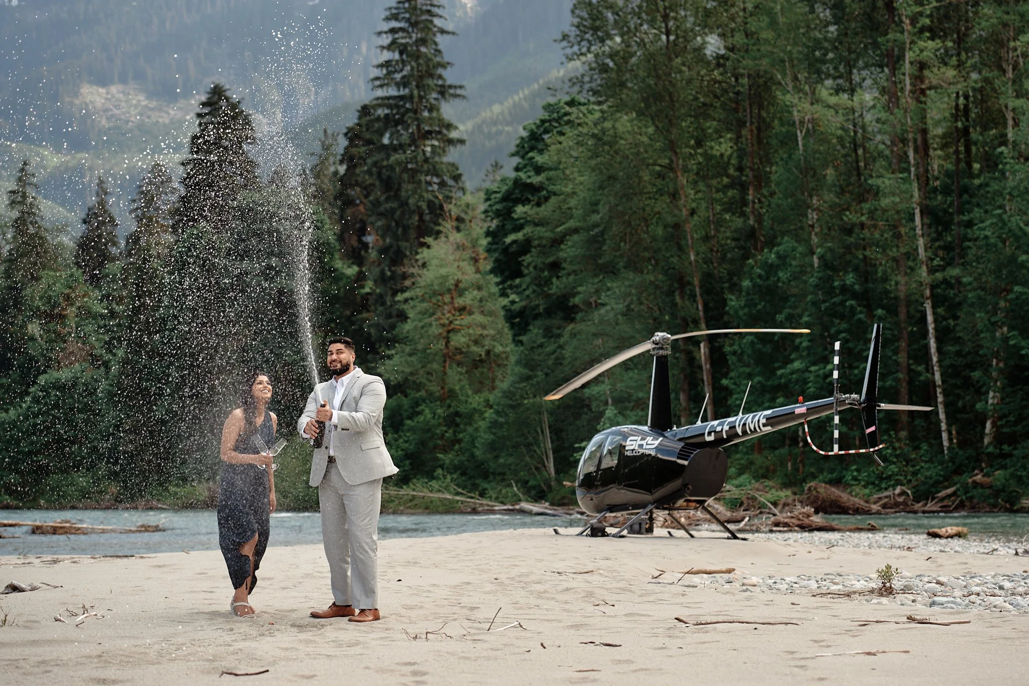 A man and woman in formal attire celebrating on a sandy riverbank, with forested mountains in the background and a helicopter nearby.