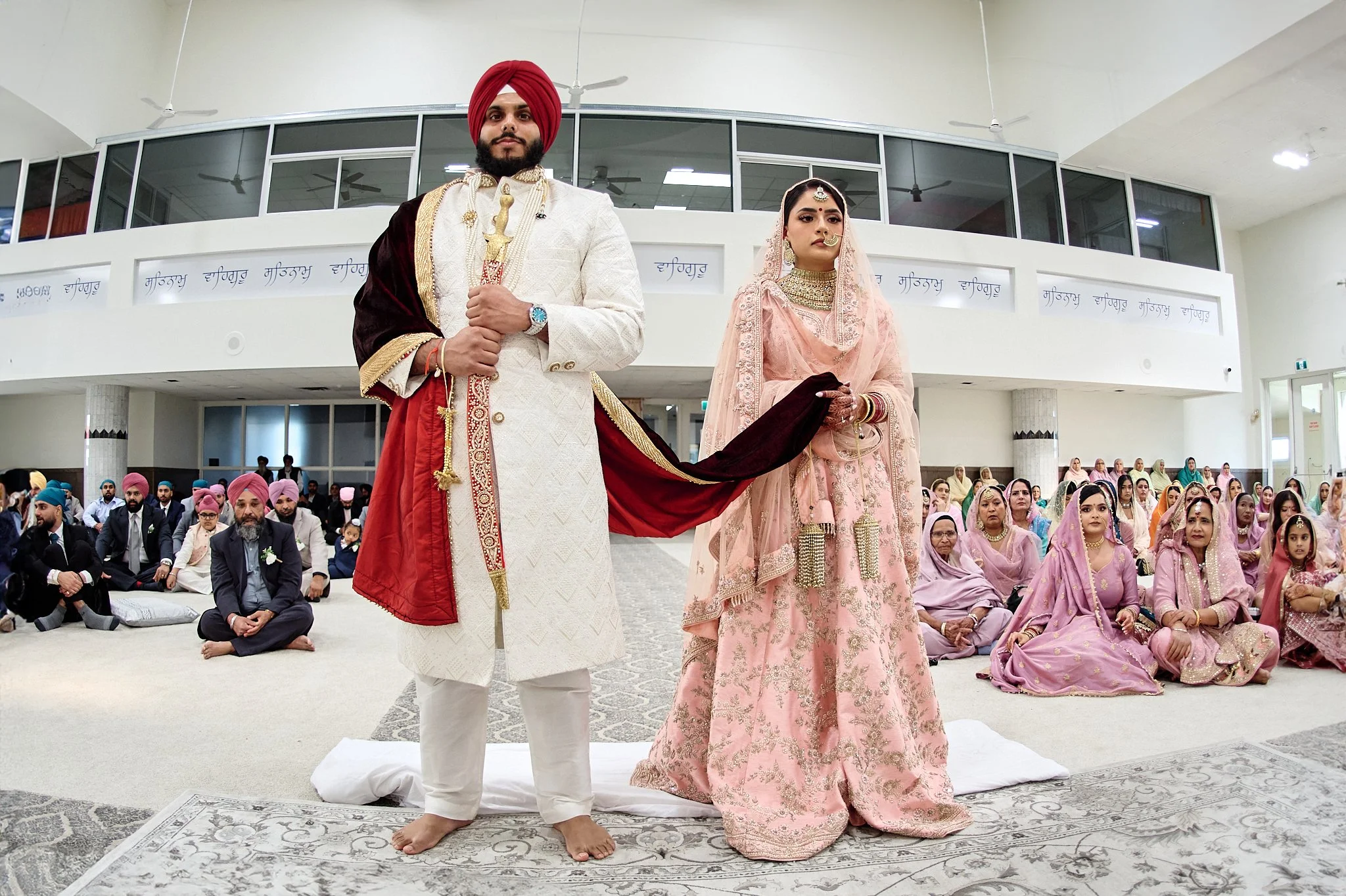 An Indian wedding ceremony with a bride and groom standing in front of seated guests. The groom wears a white sherwani with a red turban and a red and gold dupatta draped over his shoulder. The bride wears a pink and gold lehenga with intricate embroidery and traditional jewelry, including a maang tikka and nose ring. The guests are dressed in traditional Indian attire, with women in sarees and men in suits and turbans, inside a spacious hall with high ceilings and large windows.