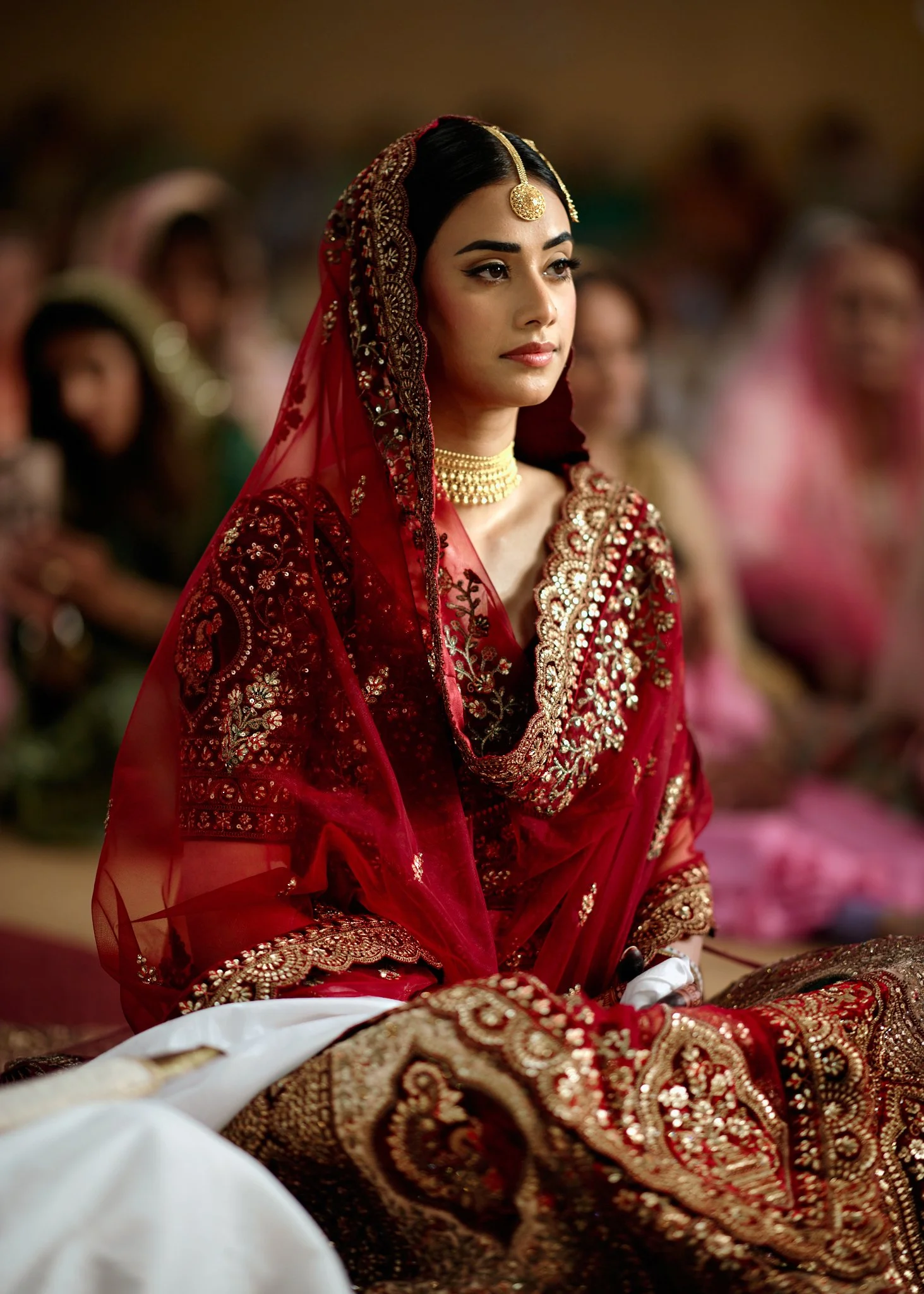A woman dressed in traditional South Asian bridal attire, wearing a red and gold embroidered outfit with a matching veil, jewelry including a choker necklace and headpiece, sitting among a group of women in similar attire.