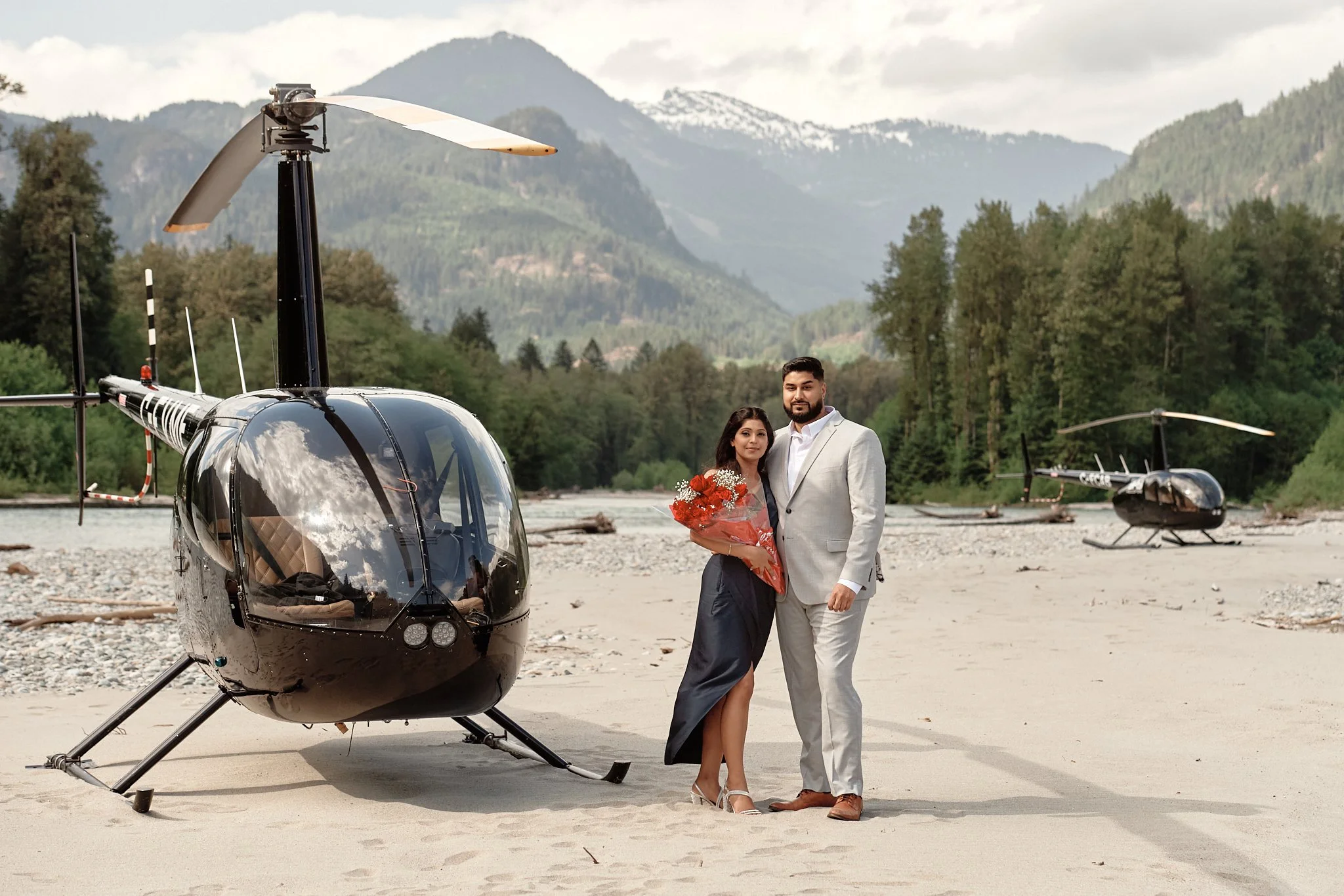 A man and a woman in formal attire standing on a sandy riverbank, holding a bouquet of flowers, with two helicopters and a mountain range with snow-capped peaks in the background.