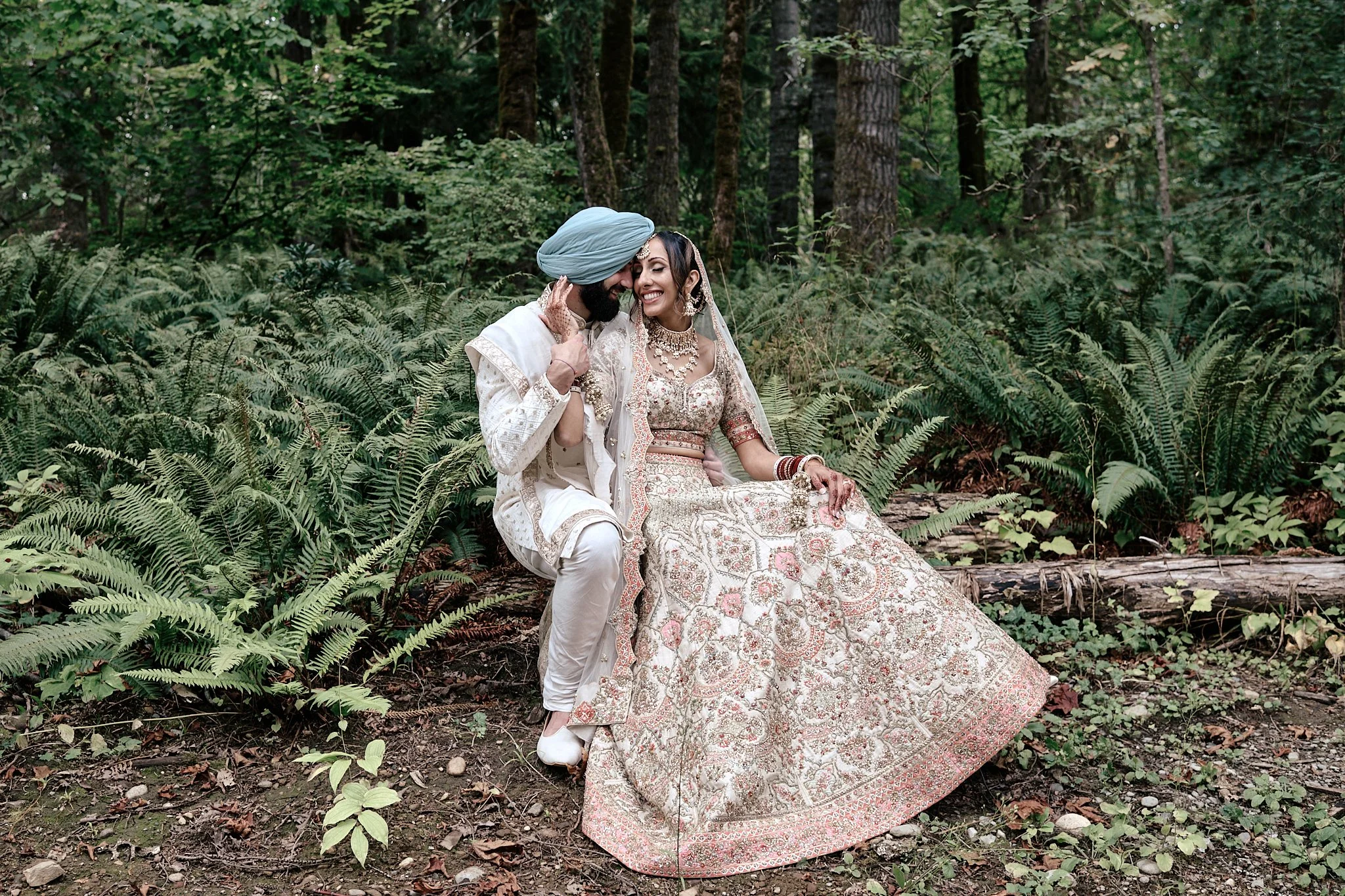 Indian bride and groom in traditional wedding attire sitting on a log in a forest, smiling at each other.