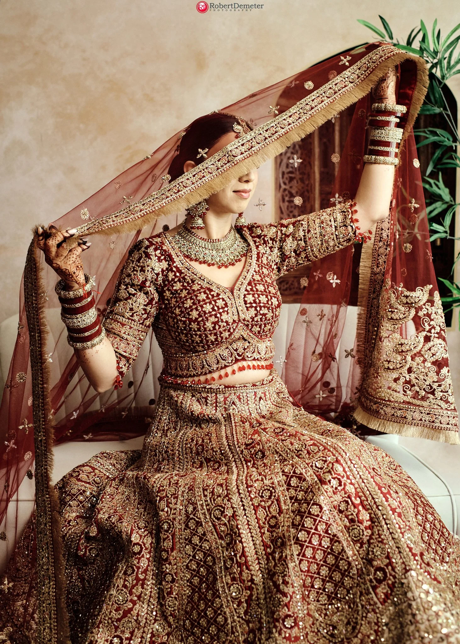 A woman dressed in traditional Indian bridal attire, wearing a heavily embroidered red and gold lehenga, with jewelry and bangles, holds a sheer red dupatta partially over her face in a decorated indoor setting.