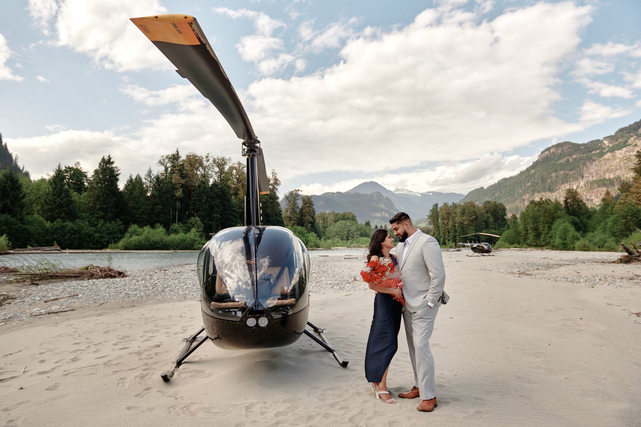 Heli engagement, a couple standing close together on a sandy riverside, holding a bouquet, with a helicopter parked nearby and a helicopter in the background, surrounded by lush trees and mountains under a partly cloudy sky. 